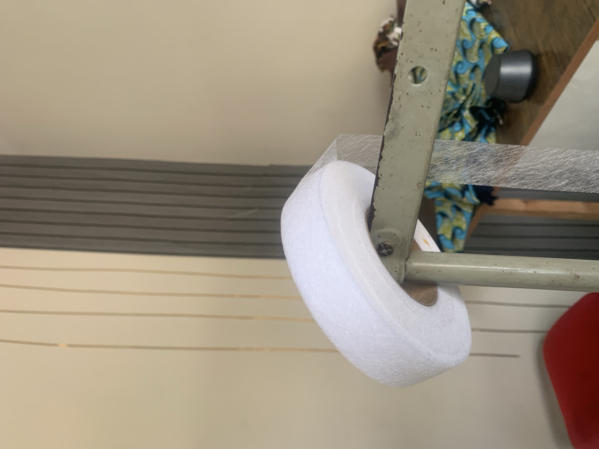 The image shows a close-up of a roll of white material, possibly foam or batting, wrapped in plastic netting. It is mounted on a metal support that appears to be part of a sewing machine or industrial equipment. In the background, a wall with vertical lines and a partially visible section of ribbed material are present. A wooden surface below holds a patterned fabric and a small black object. The scene suggests an indoor setting, possibly a workshop or tailoring space in Jauro Yinu, Nigeria.