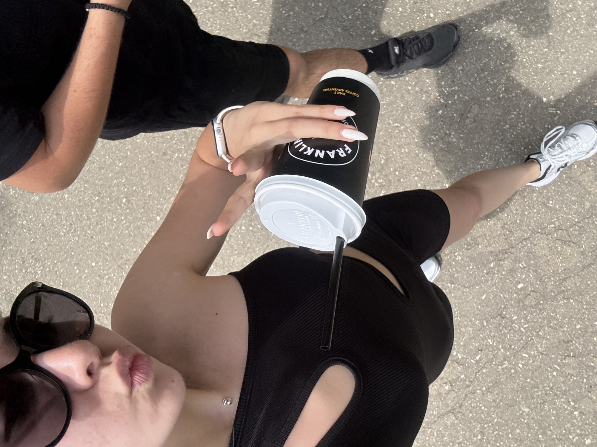 A medium shot, from the perspective of someone standing, shows two people on a paved surface. A person in the foreground, facing to the left, wears sunglasses, a black sleeveless top, and black biker shorts. They are holding a black and white Franklin brand coffee cup with a straw. The person is wearing a silver watch. A second person, partially visible, wears black shorts, a black shirt, and black athletic shoes. The location cannot be determined from the image.