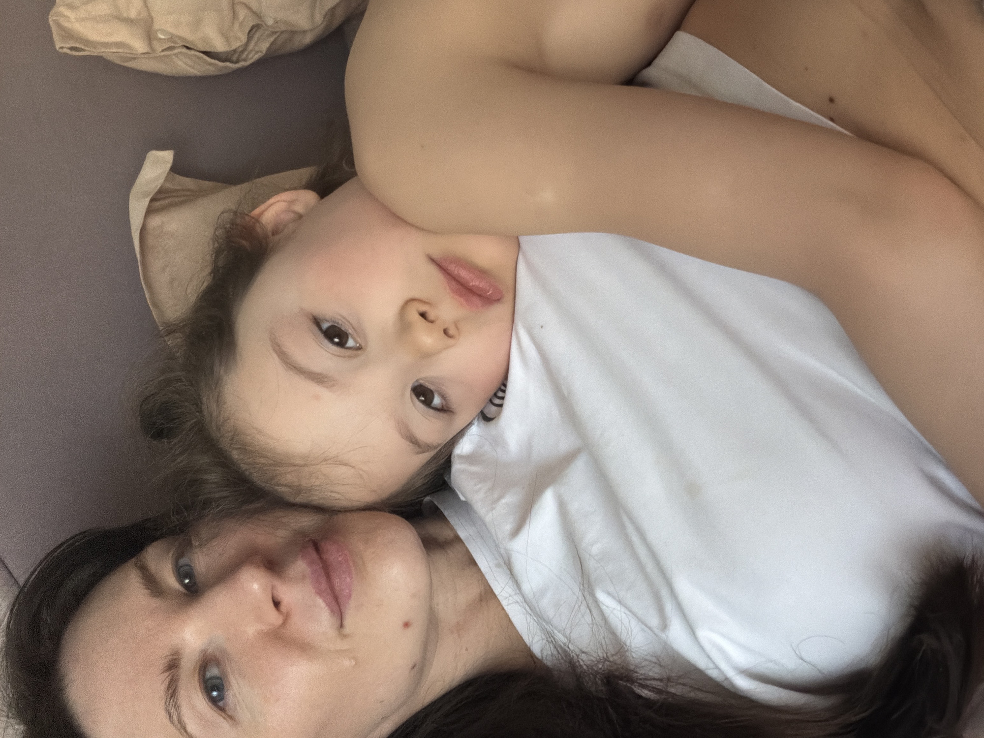 An intimate, high-angle portrait captures an adult and a child lying closely together, facing upwards towards the camera. The image is oriented such that their bodies appear rotated, with their heads positioned towards the bottom-center and their bodies extending upwards and to the right.
The adult, with long dark hair, is positioned beneath the child, her face visible in the lower left portion of the frame. She has blue eyes and a calm, slightly serene expression, with her lips slightly parted. A small, clear tear or droplet is visible on her right cheek, just below her eye.
The child, appearing younger, is lying on top of the adult, resting her head near the adult's shoulder or chest area. She has dark eyes and a neutral expression, looking directly at the camera. Her lips are slightly parted. She is wearing a plain white t-shirt, which contrasts with the adult's darker clothing (partially visible). One of the child's arms is draped over the adult's body, and further down, the child's bare skin (arm and part of a leg) and more white fabric are visible, suggesting light clothing or a blanket.
The setting appears to be indoors, likely on a soft surface such as a bed or sofa. A crumpled, light beige fabric object (possibly a pillow or blanket) is visible in the upper left corner of the frame, and a muted grey fabric surface forms the background beneath the adult. The lighting is soft and even, suggesting a relaxed atmosphere. The overall impression is one of warmth, affection, and close bond between the two individuals. There is no visible text in the image.
