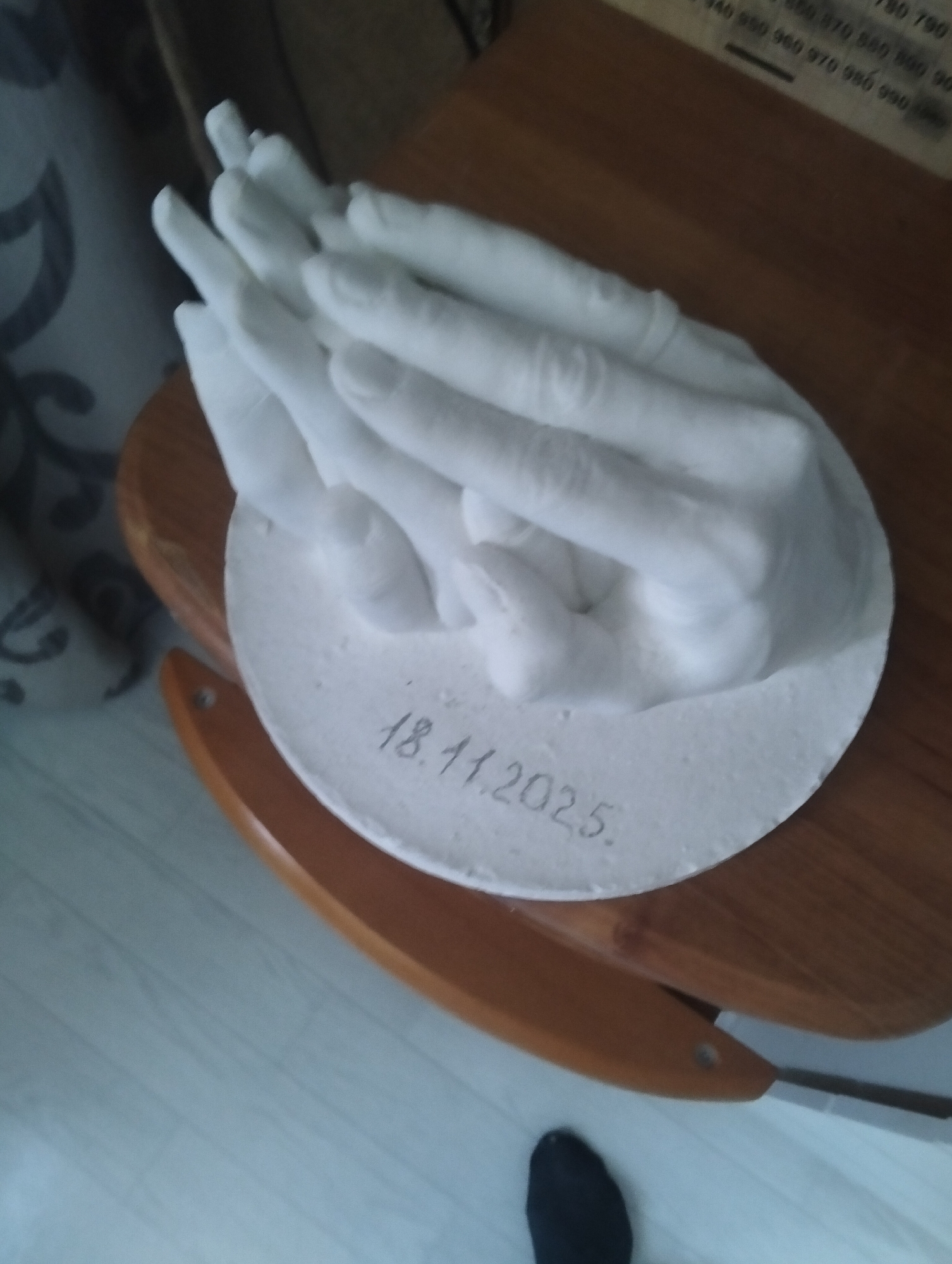 The image shows a plaster cast of a pair of hands. The cast is on a circular white base, with the date "18.11.2025" inscribed on it. The hands appear to be intertwined. The cast is placed on a wooden surface, possibly a table or a piece of furniture. In the background, there is a hint of a patterned fabric. The lighting suggests an indoor environment. The image does not provide any specific visual cues to determine its location in Cherkessk, Russia.