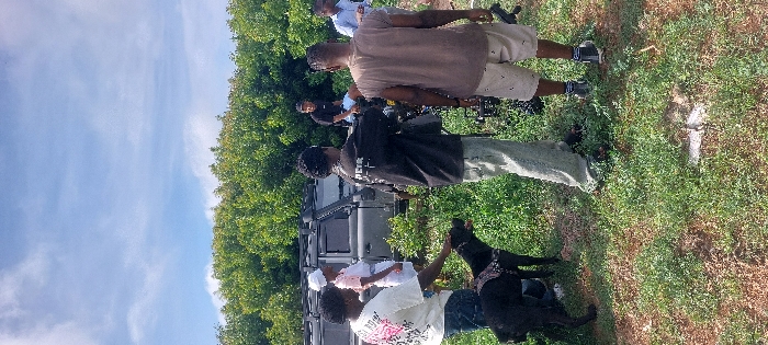 The image depicts an outdoor scene under a partly cloudy blue sky, characteristic of a sunny day, located in Dar es-Salaam, Tanzania. The specific location within the city cannot be determined from visual cues.
In the foreground and midground, a group of six individuals and one dog are present on uneven ground covered with sparse green grass and visible reddish-brown soil. A dense line of green trees forms the background.
From left to right:
1. A person, possibly female, wearing a white t-shirt with a graphic design and blue jeans, is seen from the side, petting a black, short-haired dog wearing a dark harness.
2. The black dog is positioned facing right, being petted by the person to its left.
3. Behind the first person and the dog, a dark-colored SUV, resembling a Land Rover Defender, is parked. It is equipped with a roof rack and a snorkel on the driver's side.
4. Near the left rear of the vehicle, a person, possibly female, wearing a white chef's hat and a white apron over a light pink top, stands facing right.
5. In the center, a person, possibly male, wearing a dark t-shirt with white text on the back and light-colored cargo pants, stands with their back mostly to the viewer, holding what appears to be a camera rig.
6. To the right of the person with the camera rig, another person, possibly male, wearing a light brown sleeveless shirt and light-colored shorts, stands with their back to the viewer, holding an object in their right hand, possibly related to camera equipment.
7. Further to the right and partially obscured, a person wearing a light blue collared shirt is visible.
8. In the background, seated on the roof rack of the SUV, is a person, possibly male, wearing a dark t-shirt and light shorts.
The people appear to be engaged in an activity, possibly a filming or photography session, given the visible camera equipment. The ground contains some scattered debris, including a light-colored object near the center of the lower frame.