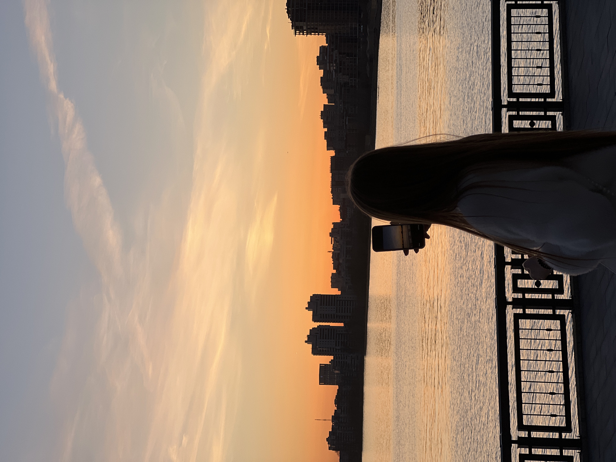 The image displays a picturesque sunset over a city waterfront, likely a promenade. In the foreground, the silhouette of a person with long, dark hair is visible from the back, holding a smartphone with both hands to capture the scene. They stand beside a dark, decorative metal railing or fence that runs along the edge of the water.
The sky is a prominent feature, displaying a warm gradient of colors typical of sunset or golden hour, transitioning from bright orange and yellow hues near the horizon to a softer blue-grey towards the top. Wispy clouds are scattered across the upper sky. Below the horizon, a calm body of water, possibly a river or lake, reflects the golden light, showing subtle ripples on its surface. Across the water, the dark silhouette of a city skyline is visible against the vibrant sky, composed of numerous buildings of varying heights, indicating an urban environment. No visible text is present in the image. The scene is set in Kazan, Russia, and evokes a tranquil and beautiful atmosphere.