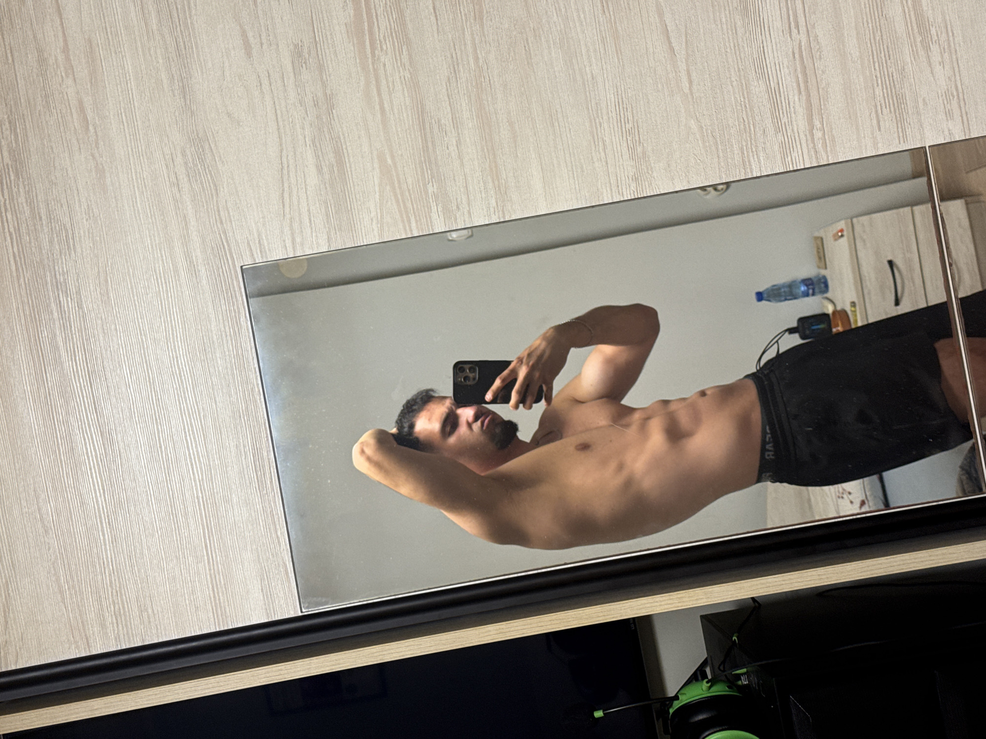 The image depicts a shirtless man with well-defined abdominal muscles posing for a selfie in a mirror. He is wearing black shorts. The man is looking at his phone, which he is holding to take the picture. The mirror reflects the man and the room he is in. The room appears to be a bedroom or dressing area, with a wooden dresser visible in the reflection. The wall behind the dresser is light-colored. To the left of the mirror, a black object with green headphones is visible, possibly a computer or gaming setup. The flooring is light-colored wood. The image is taken from a high angle, looking down at the man and the mirror. There is no indication of the location being in the city of Shumen, Bulgaria.