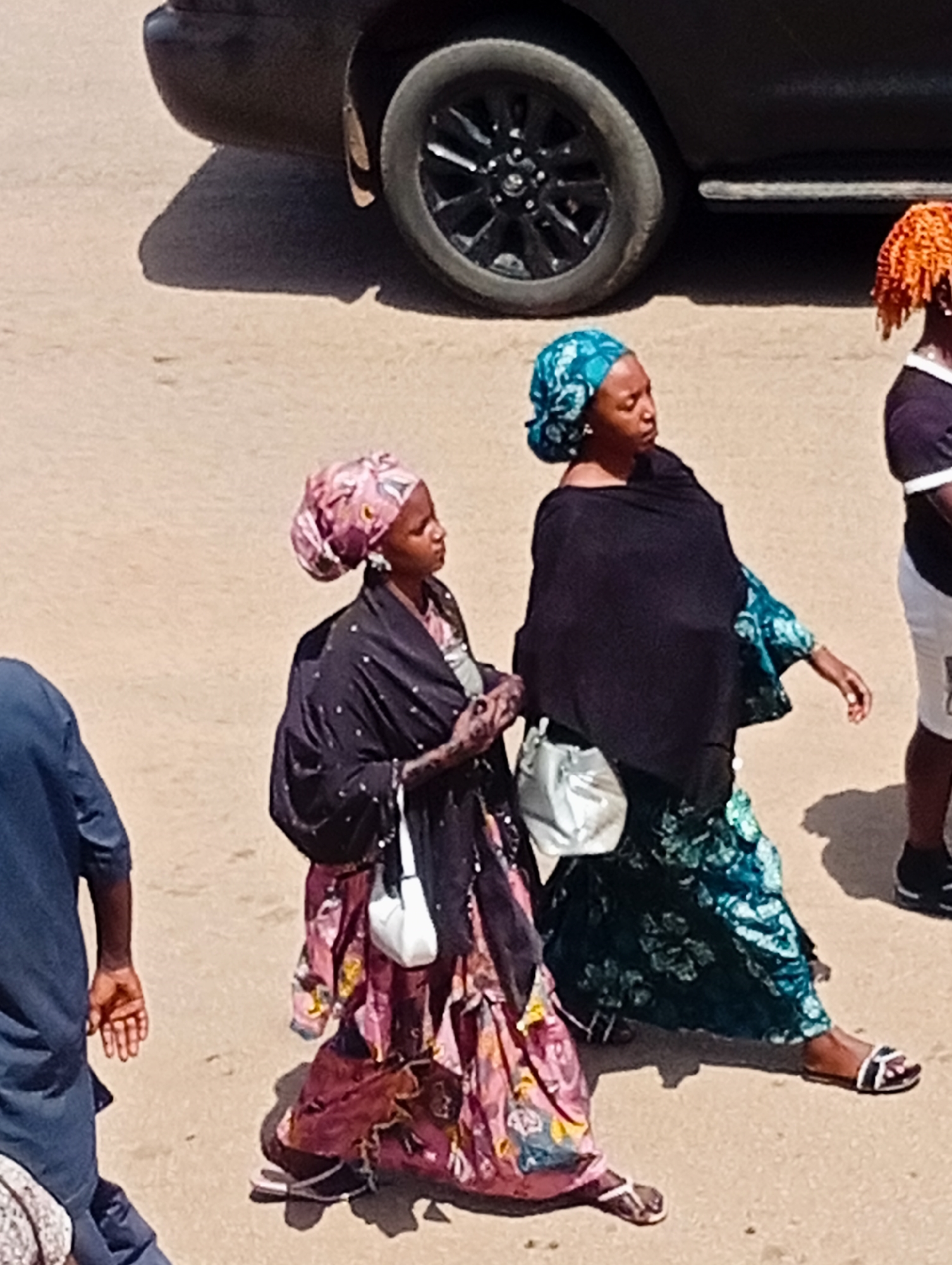 Two women in traditional attire walk on a dusty street in Bukuru, Nigeria. The woman on the left wears a pink and patterned headscarf, a dark patterned djellaba, and a colorful skirt. She carries a small white purse. The woman on the right wears a teal headscarf and a dark shawl over a patterned green dress. She also carries a white purse. They are walking away from the camera, towards the right of the frame. In the background, a dark SUV is partially visible, with its rear wheel and part of the bodywork showing. The ground is sandy and appears to be a dirt road. Sunlight casts shadows on the ground.