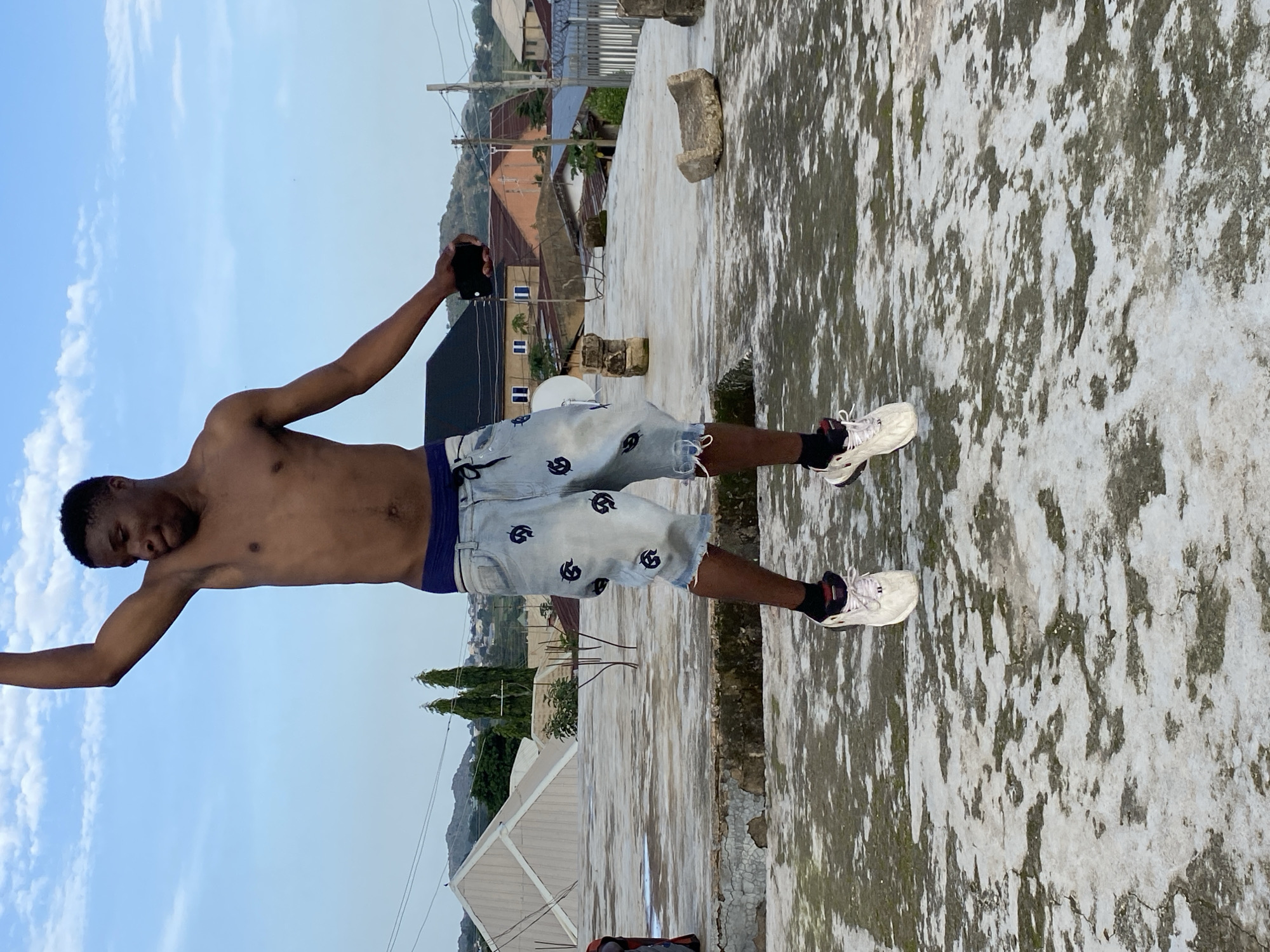 A young man, shirtless and wearing light-colored denim shorts with a dark pattern, stands on a rooftop with a cell phone in his right hand, appearing to take a photo or video. He is wearing white athletic shoes with black socks. The rooftop is weathered with a mottled grey and white surface. In the background, there are houses with corrugated metal roofs and some vegetation, suggesting a residential area. The sky is a clear blue with a few scattered white clouds, indicating it is daytime. The overall scene is casual and relaxed, with the man posing for a photograph. The location context suggests this is Bukuru, Nigeria.
