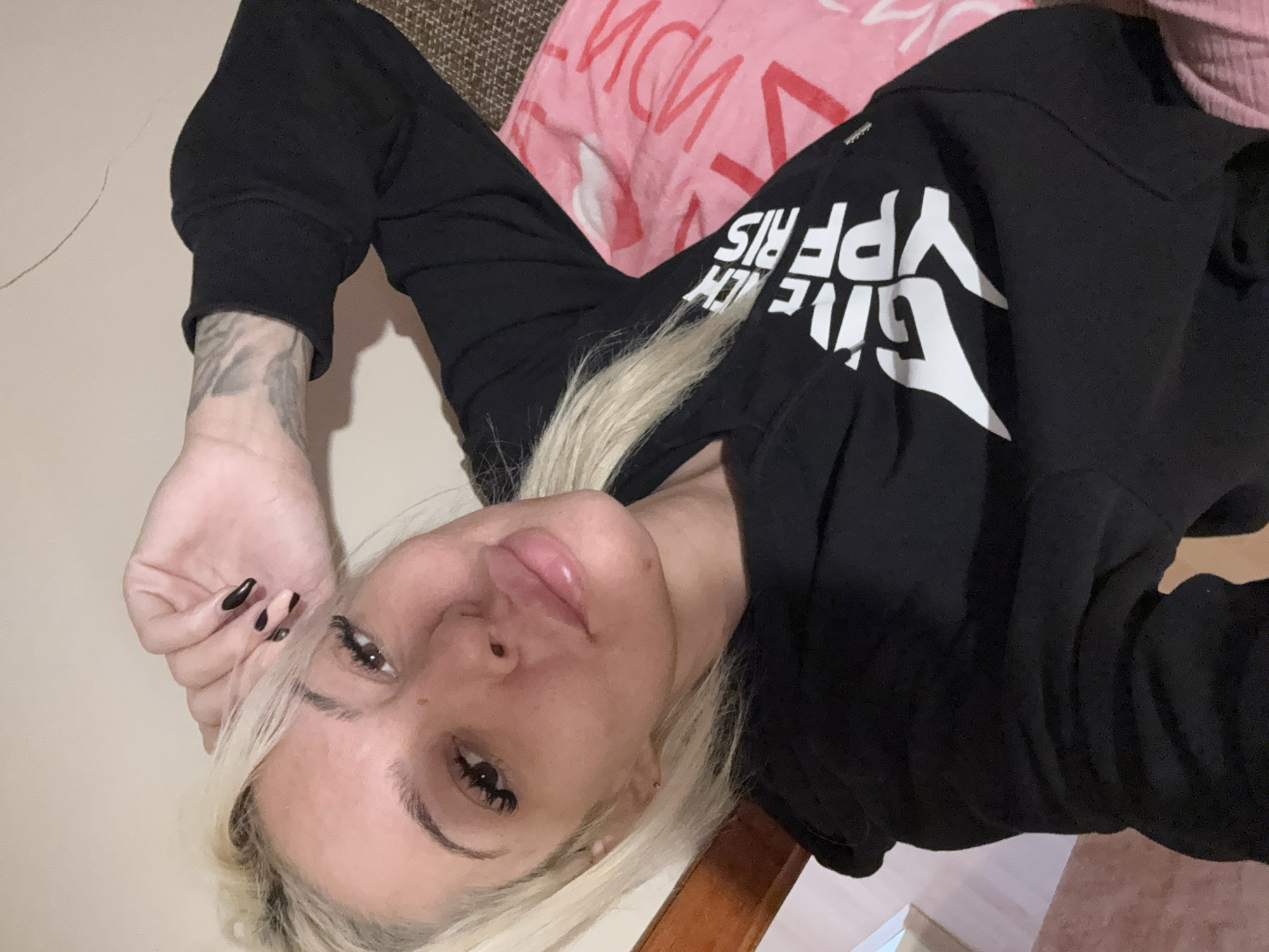 A young woman with blonde hair and fair skin is captured in a close-up portrait, seemingly a selfie, as she lies or reclines indoors. Her head is tilted, and she looks directly at the camera with a calm, neutral expression. She has dark, defined eyebrows, long eyelashes, and full, glossy lips.
She is wearing a black long-sleeved top or sweatshirt with white text on the front that clearly reads "GIVENCHY PARIS". Her right wrist displays a tattoo featuring light grey, abstract, or feathery patterns. Her left hand, with nails painted in a black-tipped, nude-based design, is visible near her face.
The background includes a plain, light-colored wall, a section of brown patterned fabric (possibly a couch or cushion), and a pink blanket or piece of fabric with some red, partially obscured lettering. A dark wooden furniture piece is also visible in the lower right corner. The indoor lighting suggests a home environment. The image itself does not contain specific visual cues to denote the geographical location of Plovdiv, Bulgaria.
