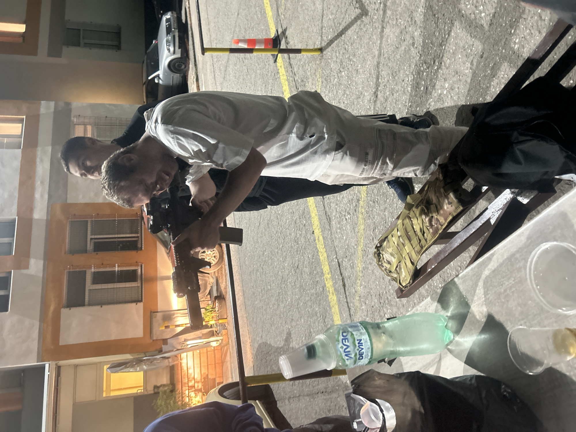 The image shows two men outdoors at night. One man is holding a firearm and appears to be aiming it. A second man stands behind him. An orange and white traffic cone is visible in the background. There is a bottle of "Devin" brand water, a table, a chair, and a few cups. The location appears to be near a multi-story building with windows and a lighted entrance, and a car is parked nearby. The building appears to be located in the city of Veliko Tarnovo, Bulgaria due to the brand of the water bottle, Devin, which is a common brand there.