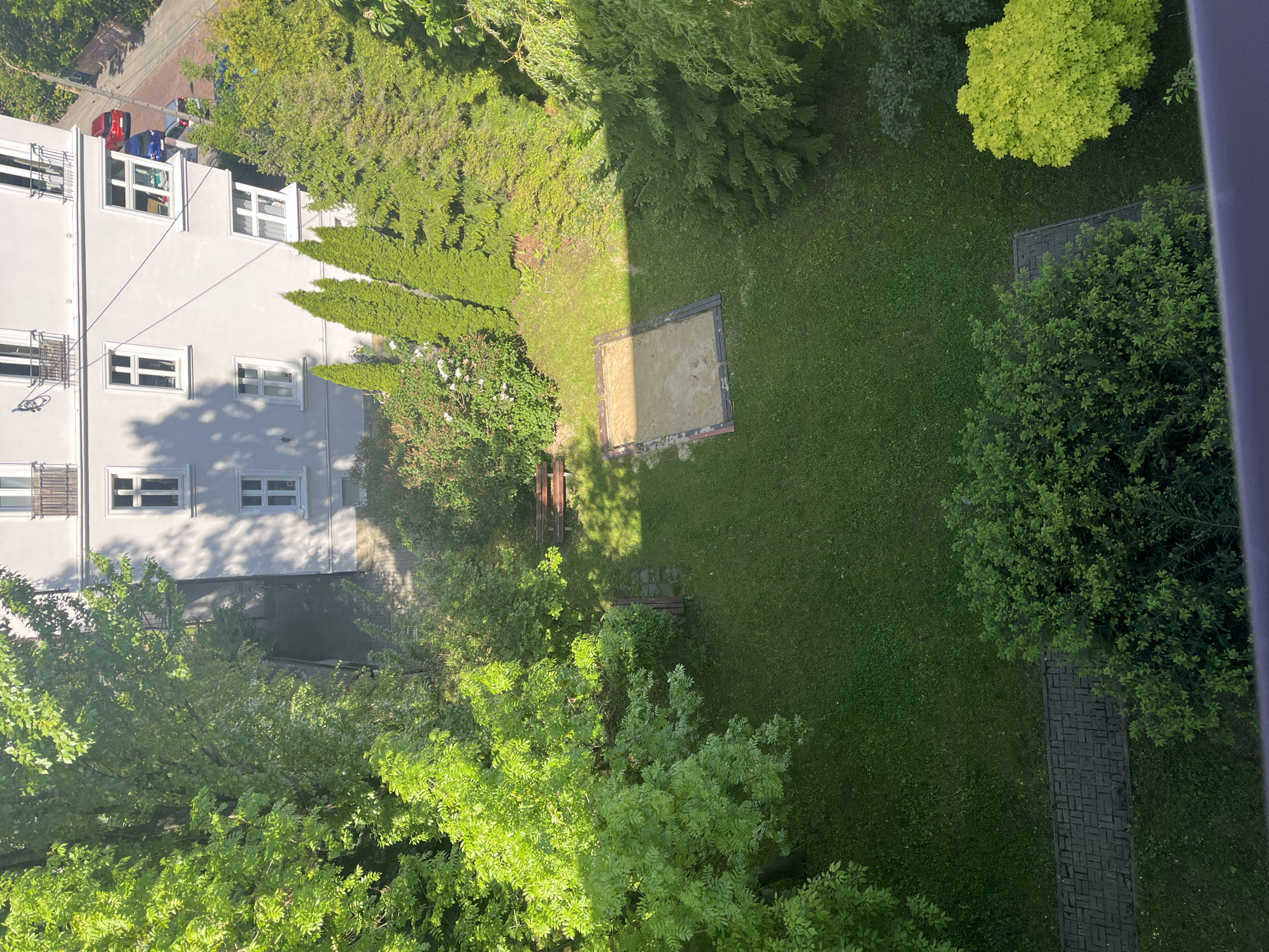 The image captures an elevated view of an urban green space located next to a multi-story building. The building is painted light grey with a series of windows, some of which have bicycles mounted on the exterior. Red and blue luggage are visible on the roof of the building. The green space comprises grass, several trees of varying species and maturity, and a rectangular sandpit. The scene is illuminated by sunlight, casting shadows across the building and the green space. There are benches near some trees. A brick path extends from the building toward the trees. The scene appears to be devoid of people.