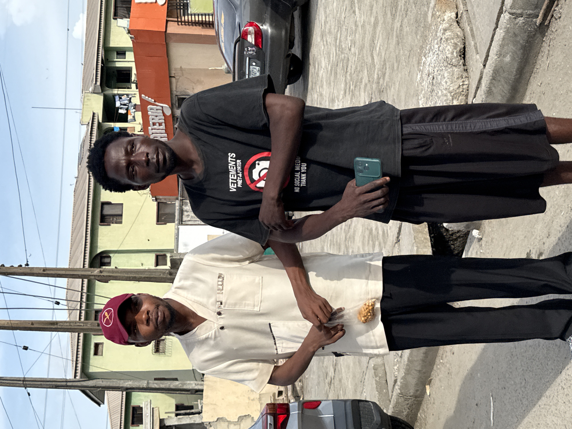 A photograph captures two men standing outdoors in a bright, urban setting, likely during the daytime under clear skies. The image is oriented vertically (portrait).
The man in the foreground on the left wears a black short-sleeved t-shirt and black shorts. His t-shirt features text: "VETEMENTS PRÊT-À-PORTER" on the left chest, and on the right side, a red circle with a diagonal line over a camera icon, accompanied by the phrase "NO SOCIAL MEDIA THANK YOU". He holds a dark green smartphone in his right hand. He has short, dark hair and a neutral expression, looking directly at the camera.
The man on the right, slightly behind the first, wears a light-colored (cream or off-white) short-sleeved shirt with dark trim details and black trousers. He is also wearing a maroon baseball cap with a small, light-colored logo featuring what appears to be two crossed objects. He holds a small, clear plastic bag containing groundnuts or peanuts in his hands. He also looks towards the camera with a neutral expression.
The background features residential buildings of varying colors, including light green and orange. Part of a word, "TERIA," is visible on the orange building, suggesting a larger word like "CRITERIA" or "NIGERIA". Utility poles with overhead wires run across the upper left of the frame. A dark gray or black car, possibly a sedan, is parked behind the men, with a partial license plate "647 JK" visible. The ground is paved, with a curb running along the right side of the image.