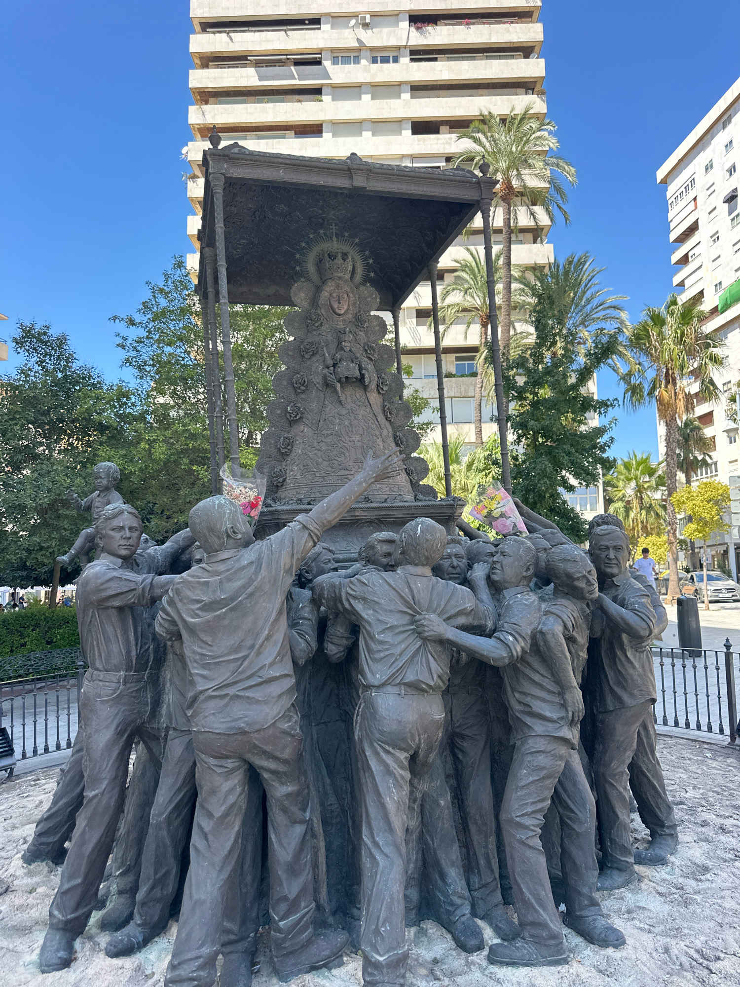 This is a medium shot, eye-level, full-color, natural light photograph of a bronze sculpture in a public square in Huelva, Spain. The sculpture depicts a group of men carrying a religious effigy of the Virgin Mary on their shoulders. The Virgin Mary is depicted with a crown and stars around her head and is adorned with roses. The men are dressed in casual clothing, with some wearing shirts and pants, and others in what appear to be work clothes. They are straining under the weight of the effigy, with expressions of effort on their faces. Some of the men are looking up at the Virgin Mary, while others are looking forward. One man in the foreground is looking directly at the camera. A child is perched on the shoulders of one of the men. A bouquet of flowers is placed on the effigy.
In the background, there are several modern buildings with balconies and palm trees. The sky is clear and blue. The sculpture is placed on a concrete base. The overall impression is one of devotion and community effort. The sculpture is located in Plaza de Las Monjas in Huelva, Spain.