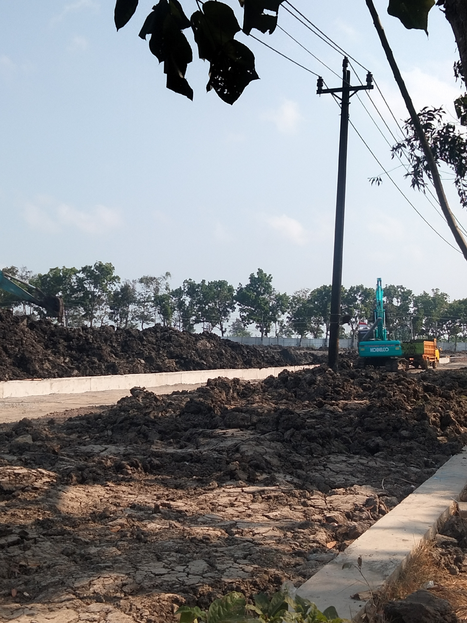 The image depicts a construction site in Sikampuh, Indonesia, with a clear blue sky overhead. In the foreground, mounds of dark soil and cracked earth dominate the scene. A light-colored concrete barrier runs along the right side of the frame, separating the soil from what appears to be a pathway. In the middle ground, an excavator, identified by the "KOBELCO" logo, is actively moving soil. To the right of the excavator, a yellow dump truck is positioned, presumably to receive the soil. Behind the construction activity, a row of lush green trees forms a backdrop against the horizon. A prominent utility pole with multiple wires stretches vertically through the image, positioned slightly to the left of the center, with dark leaves from an overhanging tree obscuring the top portion of the frame. The overall scene suggests ongoing earthmoving operations at a construction location.