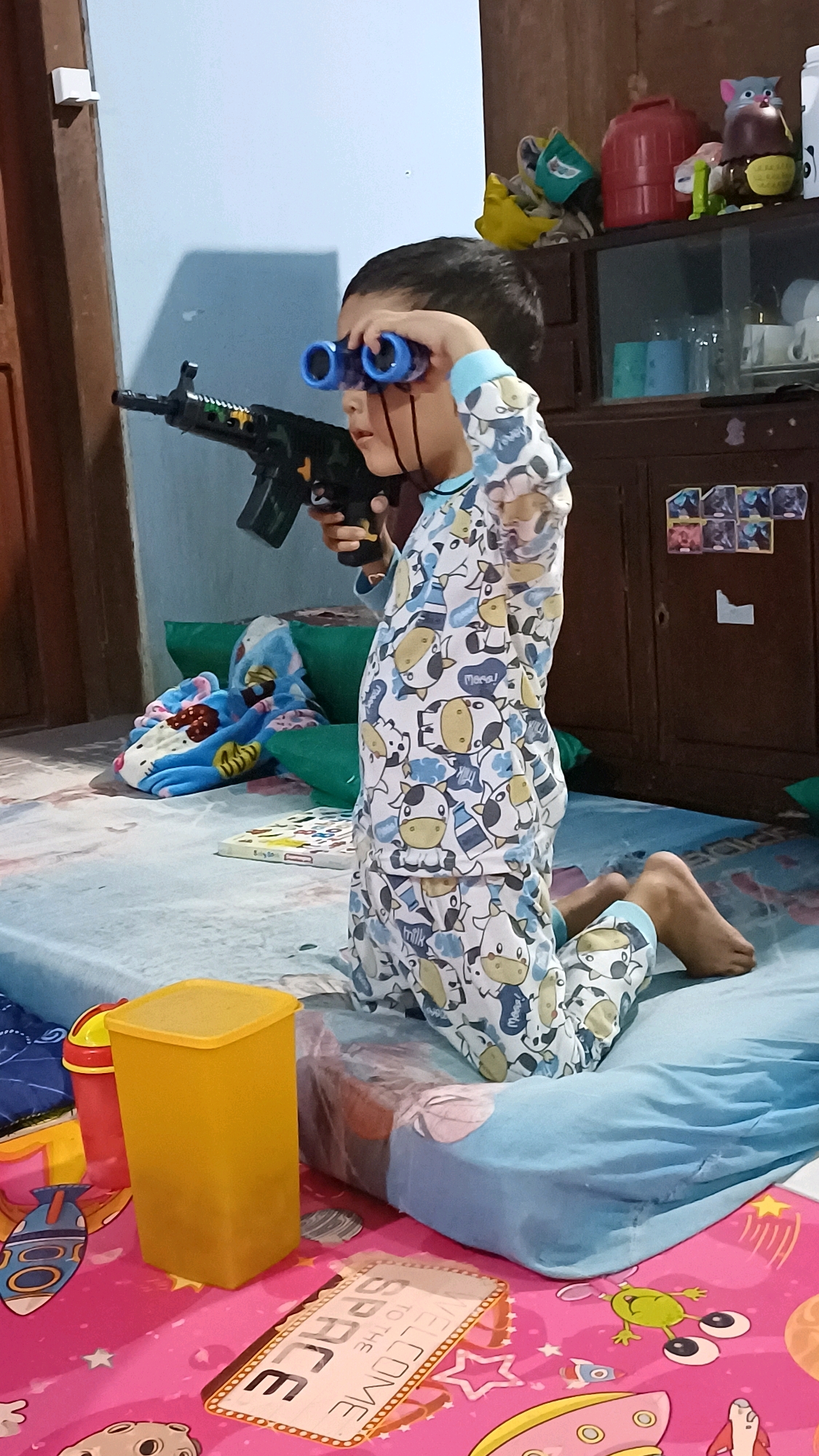 A young child is kneeling on a light blue mattress in what appears to be a bedroom in Wonokerso, Indonesia. The child is wearing light blue pajamas with a cow print and is holding a black toy rifle with a camouflaged pattern in their right hand, while also holding blue toy binoculars to their eyes with their left hand. The child's gaze is directed forward, as if looking through the binoculars. The mattress is situated on a pink floor mat decorated with cartoon rockets, stars, and planets. To the left of the child is a tall, yellow plastic container. Behind the child, several green pillows and a blue blanket with a cartoon print are visible. In the background, a wooden cabinet with shelves displays various items, including what appears to be a red thermos, a toy cat, and drinking glasses. A white light fixture is mounted on the wall to the left of the frame. The scene suggests a child engaged in imaginative play.