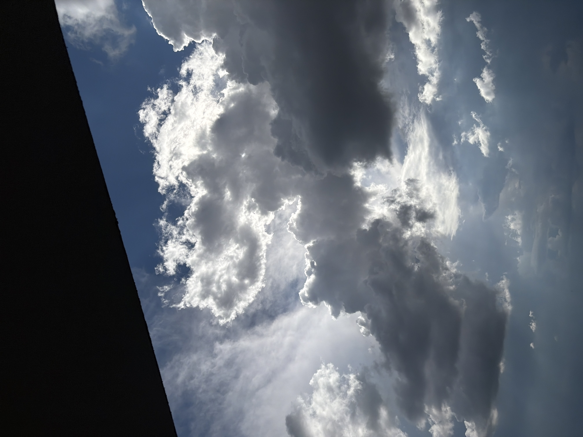 The image shows a view of the sky with clouds, taken from an unknown location in the city of Unknown, Unknown. A dark, angular structure is visible in the bottom left corner of the frame, possibly a building. The sky is a mix of blue and gray, with large, puffy clouds of varying shades of white and gray. Some of the clouds are backlit by the sun, creating a bright, ethereal effect. The overall scene suggests a dynamic weather pattern.