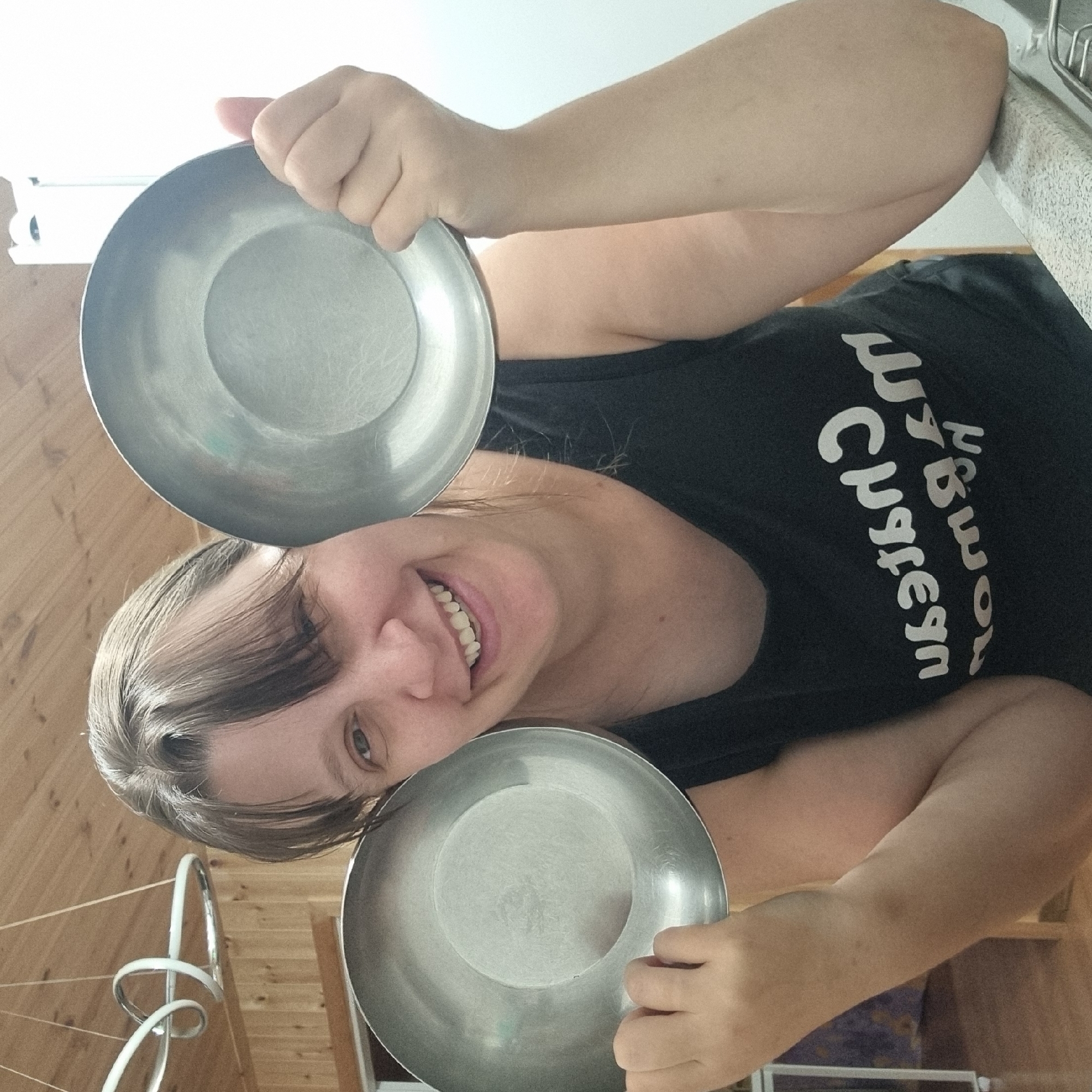 An outdoor portrait shows a woman in Dunnae-myeon, South Korea, holding two metal bowls in front of her. She is smiling broadly, with her mouth open to reveal her teeth. The woman is wearing a black tank top with white text that reads "Chateau Marmont" in a mirrored font. She has light brown hair pulled back from her face, with bangs. The background includes a wooden ceiling, a modern light fixture, and a white wall. The woman's arms and hands are visible as she holds the bowls.