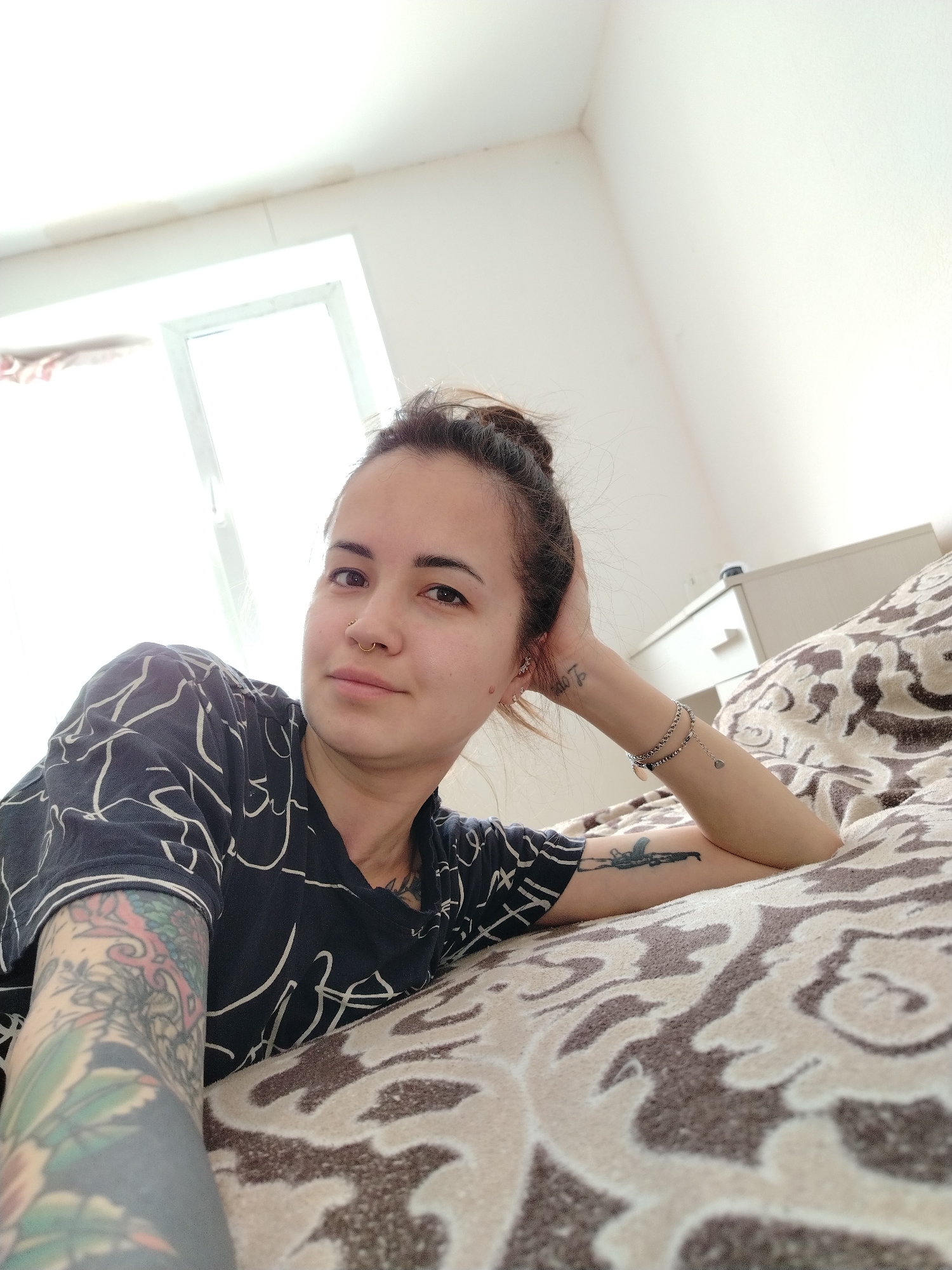 A woman lies on a bed with a patterned beige and brown blanket in a brightly lit room. The woman has her hair in a bun, a nose ring, and tattoos on her arms. She is wearing a dark t-shirt with white geometric patterns. A window is visible in the background and a white cabinet is partially visible near the head of the bed.
There is no indication of the city of Noviy, Russia.
