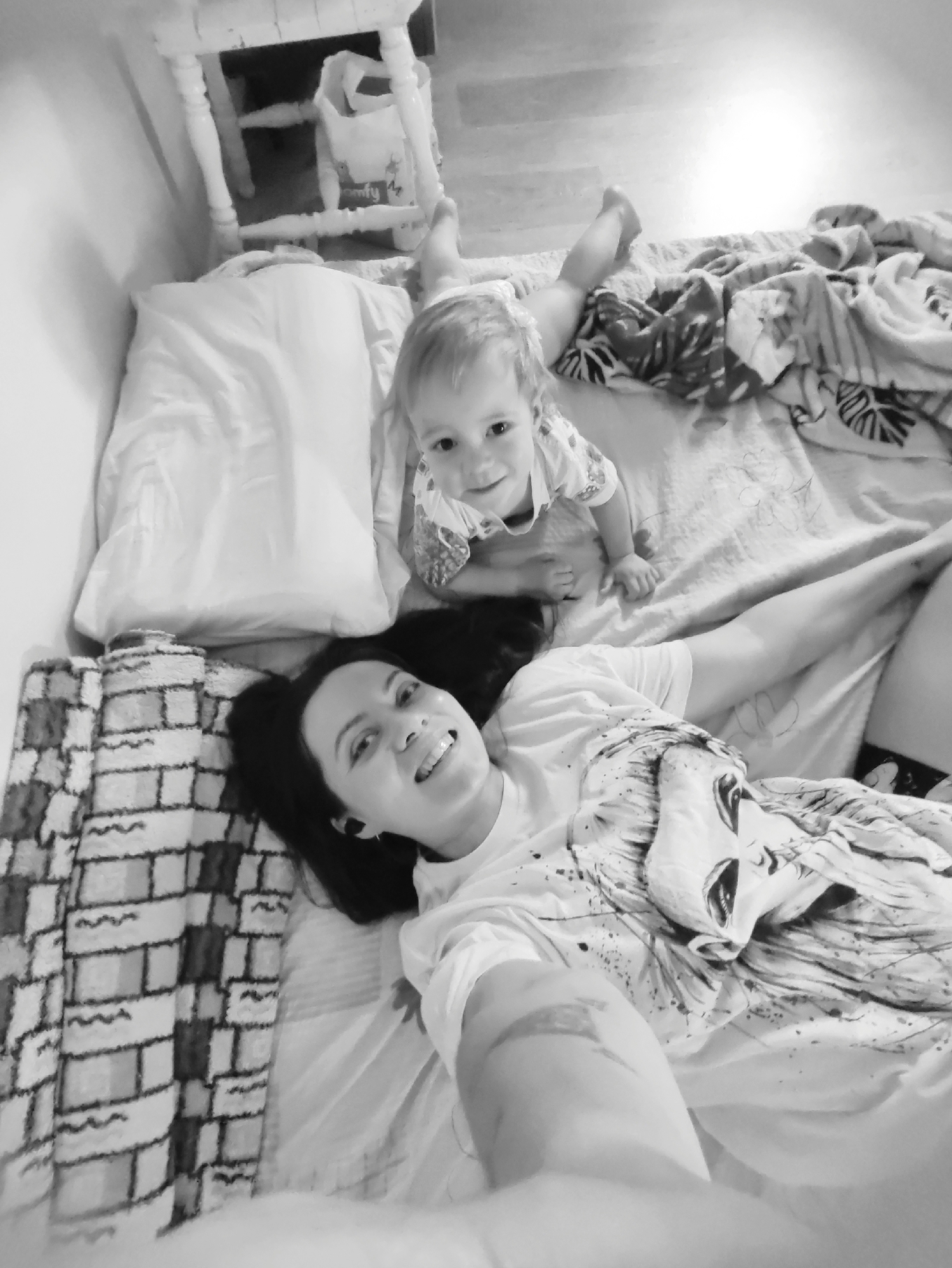 A monochrome, high-angle shot shows a woman lying on a bed with a baby. The woman has dark hair, is wearing a t-shirt with a face graphic on it, and is smiling at the camera. Her left arm is extended. The baby, located near the pillow, is crawling and also looking at the camera. A white pillow is to the woman's left, and a patterned blanket is draped over the edge of the bed. A small white table or chair is in the background. The scene's location is in the city of Unknown, Unknown.