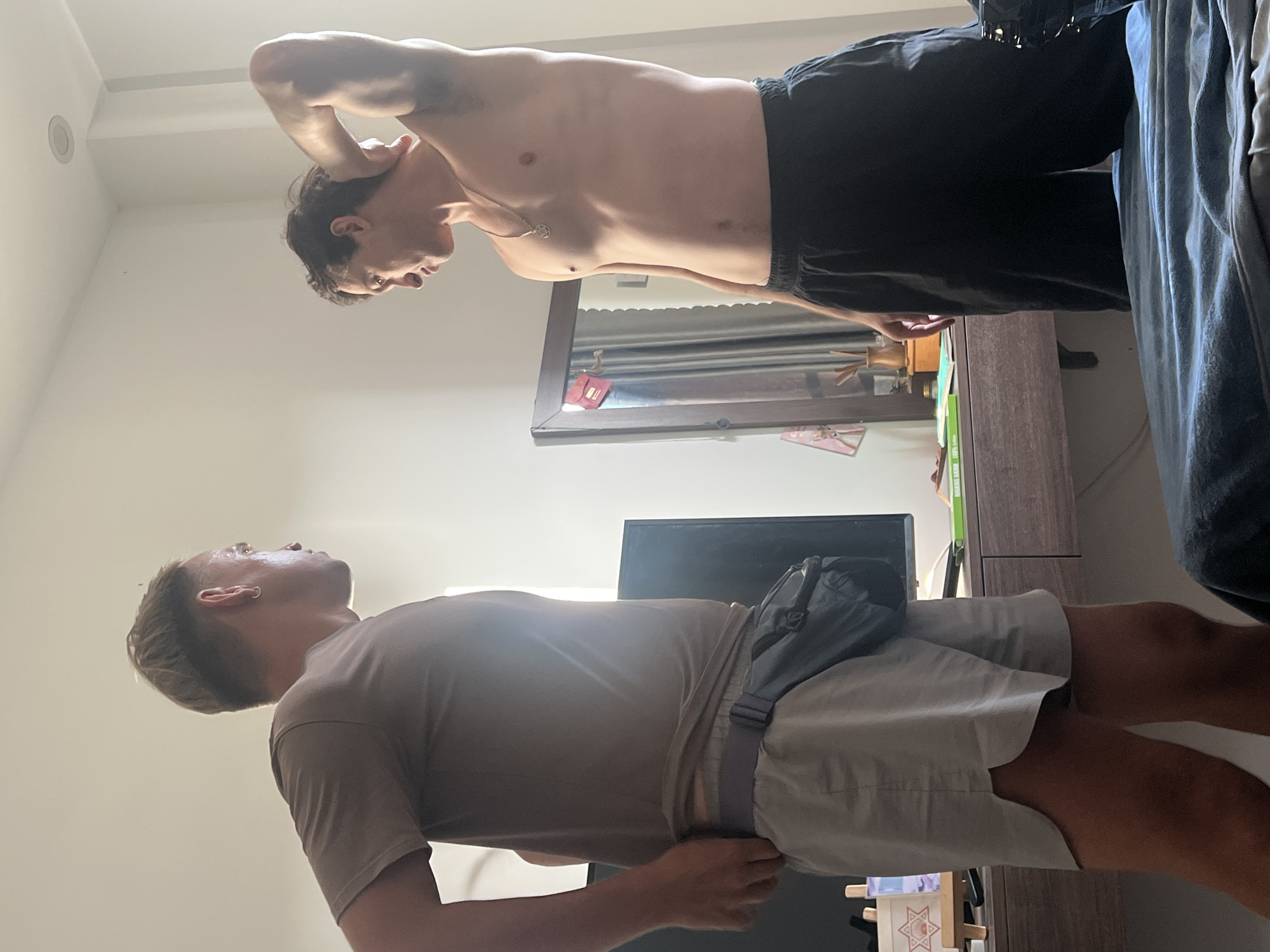 Two young men are in a room in Da Nang, Vietnam, possibly a hotel room. The man on the left is wearing a tan t-shirt and grey shorts with a fanny pack around his waist. He is facing the man on the right and appears to be adjusting his clothing. The man on the right is shirtless, wearing black shorts, and has his arm behind his head. A mirror with some decorative items is visible behind him. A desk with a television and other items is in the background. The room is brightly lit by natural light.