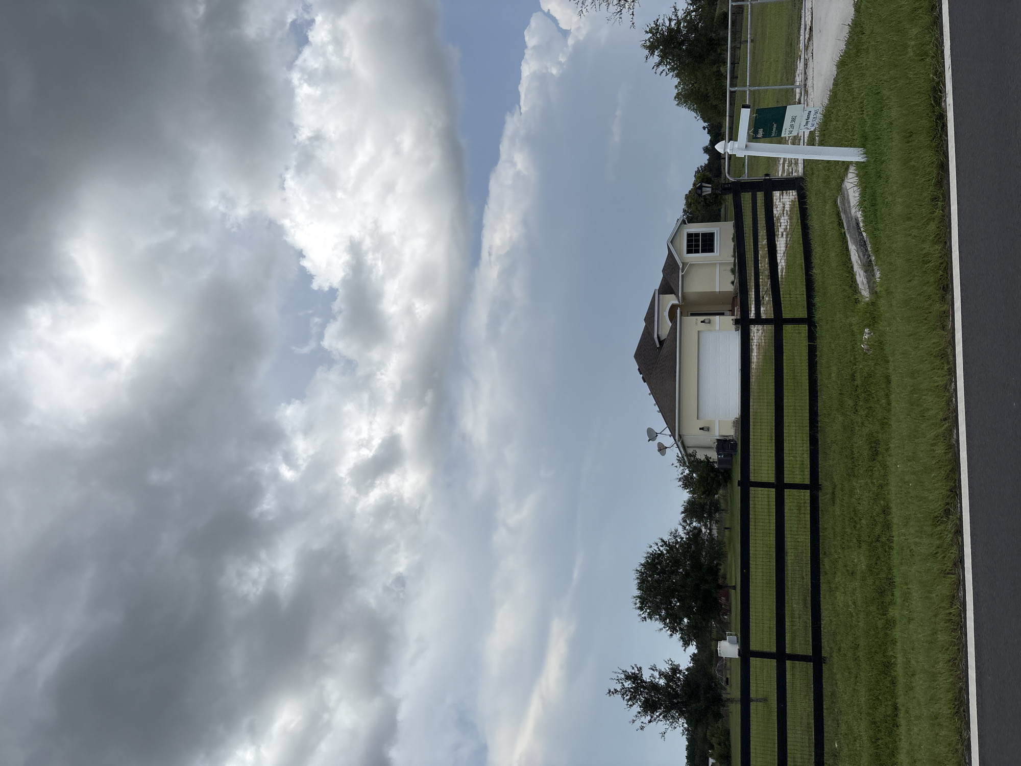 The image shows a yellow house with a white garage door and a black metal fence in front of it. The house is located in Parrish, United States, and there is a "Keyes" real estate sign in front of the fence. The sky is cloudy and there are trees in the background. The house appears to be a single-family residence. The scene is set on a grassy area with a paved road in the foreground. The overall impression is of a suburban or rural residential property.