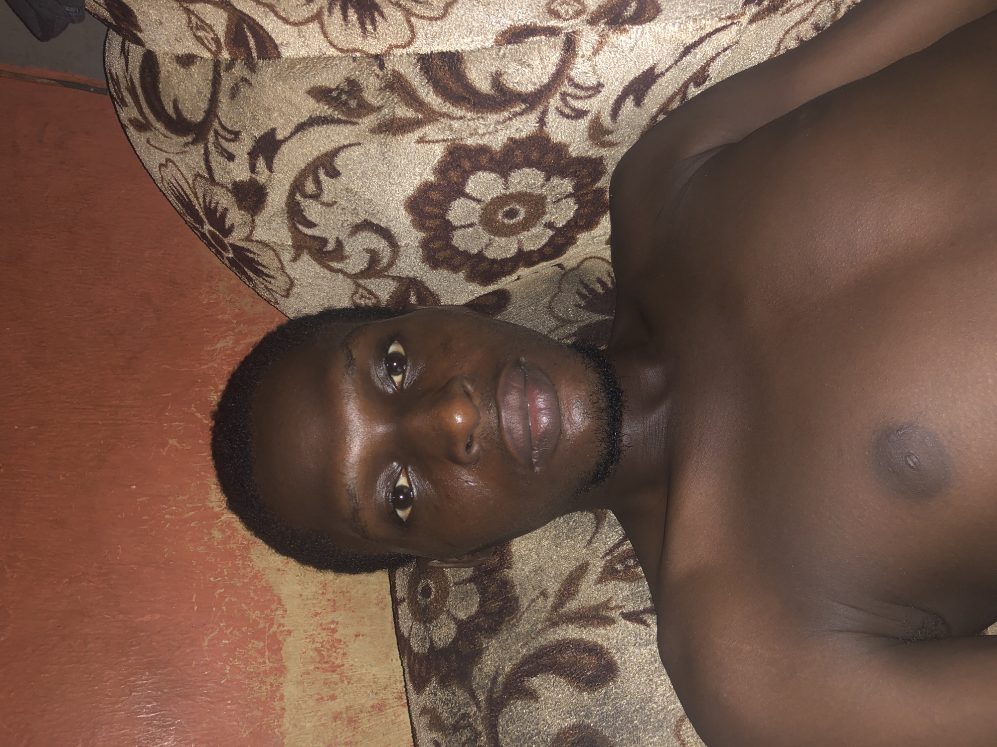 A close-up, overhead shot shows a shirtless, dark-skinned man lying on a patterned couch. The man's face is the focal point; he has short, dark hair and a neatly trimmed beard. His eyes are open and he appears to be looking at the camera. The couch is covered in a beige fabric with brown floral patterns. A section of what appears to be a painted, orange wall is visible to the side of the couch. The location is Unknown, Unknown.