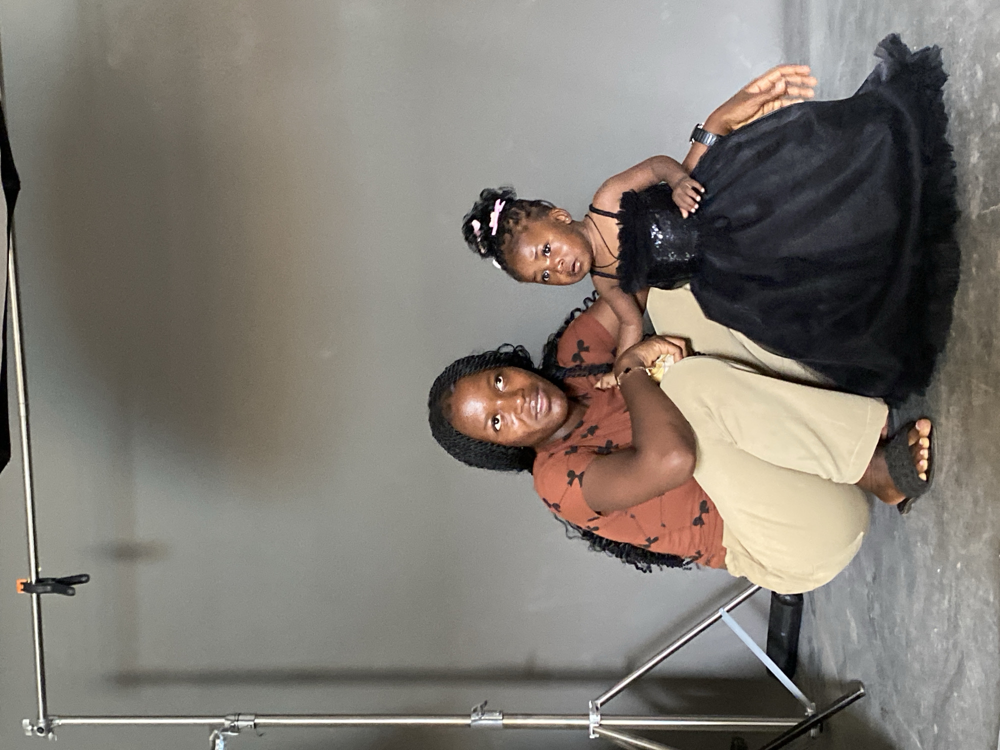 A woman and a young child are in a photography studio. The woman is crouching on a metal stand, wearing a brown patterned shirt and beige pants, and has braided hair. The child, dressed in a black dress, is positioned near the woman. The studio has a grey backdrop and the lighting is diffused. The location in the city of Jos, Nigeria, cannot be confirmed from the image.