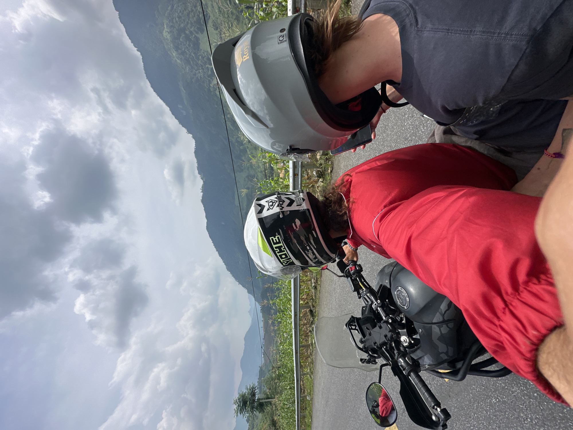 The image displays two individuals stopped with a motorcycle on a mountainous road in Minh Sơn Commune, Vietnam, under a cloudy sky. The original image is rotated, presenting the scene as if viewed from above and to the side, with the horizon running diagonally.
In the foreground, a dark grey motorcycle, possibly an adventure bike with a camouflage-patterned tank, is parked on an asphalt road. One person, wearing a red long-sleeved shirt and a white motorcycle helmet with "YOHE" and black/yellow accents, is positioned near the motorcycle's handlebars. Another individual, dressed in a dark grey t-shirt and a light grey helmet bearing the word "Royal" and an emblem, stands beside them, holding a smartphone. Both individuals appear to be looking downwards or at their phones.
A metal guardrail runs along the edge of the road. Beyond the guardrail, rows of green crops, possibly corn, are visible, cultivated on the slopes. The background is dominated by large, rolling green mountains extending into the distance under an overcast sky filled with prominent white and grey clouds. The scene suggests a break during a motorcycle journey through a scenic, mountainous landscape.