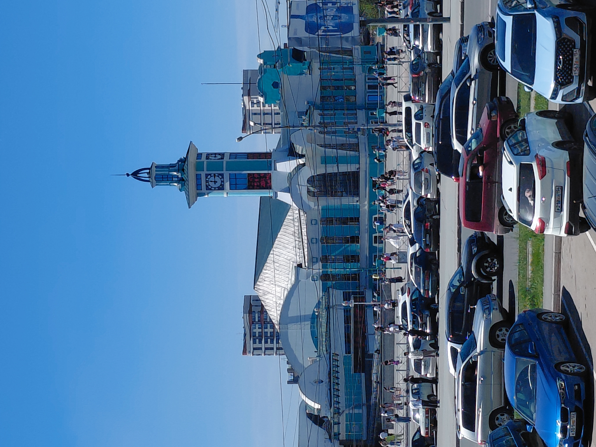 The image depicts a clear, sunny day in Novosibirsk, Russia, likely in front of the Novosibirsk Glavny railway station. In the foreground, a parking lot is filled with a variety of vehicles, including sedans and SUVs. Several people are visible walking and standing around the parking area and on a raised walkway.
Dominating the background is the impressive architecture of the railway station. It features a prominent clock tower with blue and white accents and a spire. The main building has a distinctive light blue and white color scheme, with a large, arched roof and numerous windows. The station appears to be a busy hub, with many people moving in and out.
The environment is urban, with power lines visible across the sky. The sky itself is a clear, vibrant blue, indicating fair weather. The overall impression is one of a bustling city scene on a pleasant day.