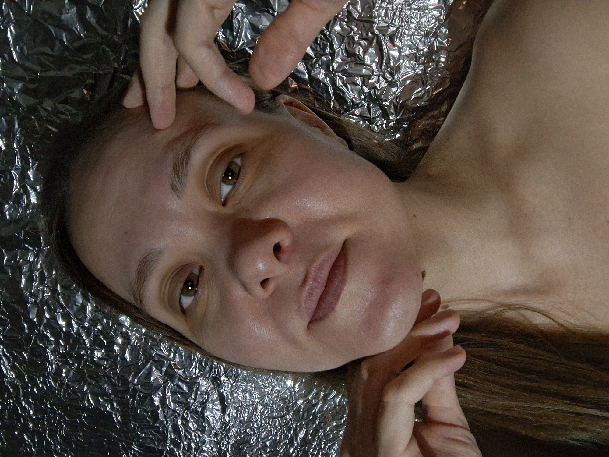 A woman lies on a crinkled sheet of aluminum foil. Her head is tilted to the left, facing the camera. Her hair is brown. Her skin is light-toned. Her eyes are brown. Her left hand is near her forehead, while her right hand is near her chin. The location is not discernible from the image. The photograph provides no indication of Chaikovsky, Russia.