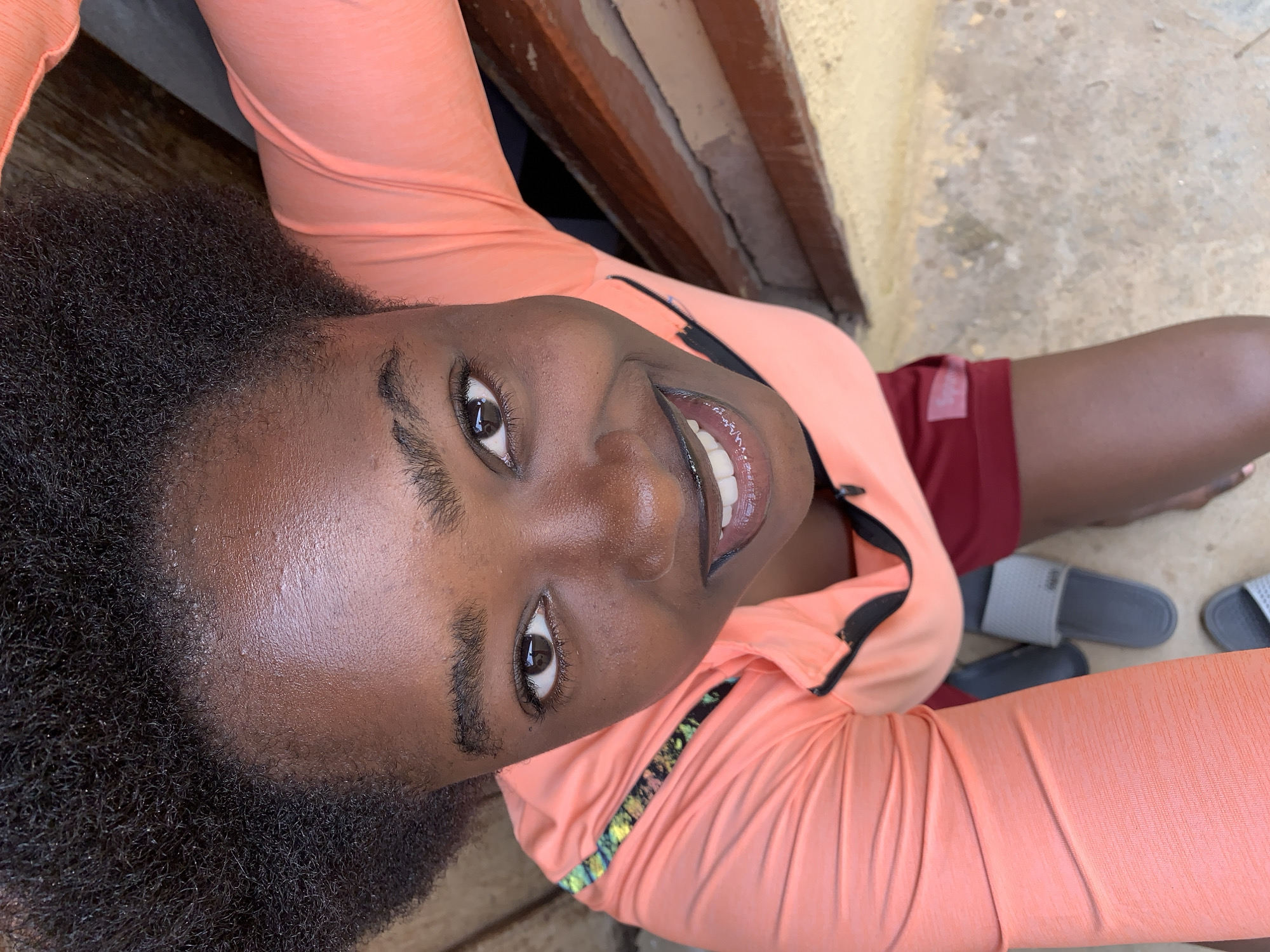 An overhead, high-angle shot captures a young Black woman from the chest up, looking up towards the camera with a wide smile, her teeth visible. She has dark, curly hair pulled back and dark brown eyes. She is wearing a coral-colored long-sleeved athletic shirt with a black zipper and a colorful striped trim along the shoulders. Below the shirt, she wears dark red athletic shorts. Her arms are raised, one of which is visible holding the camera. To her right, partially visible, is a wooden structure. On the ground below her are a pair of gray slip-on sandals. The scene is outdoors, on a concrete or dirt surface. The lighting suggests it is daytime. This scene could be located in Rafin Par, Nigeria.