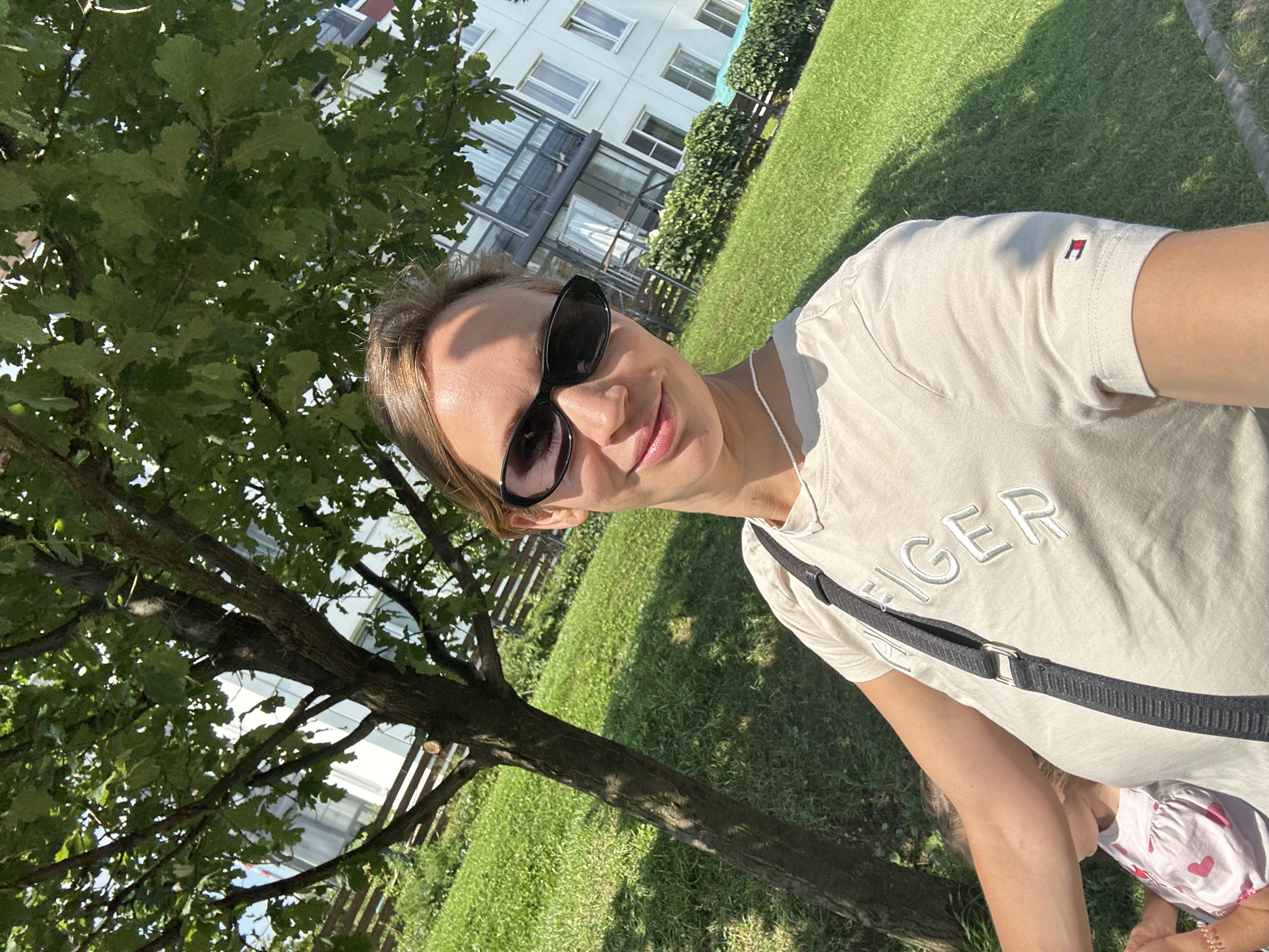 In Vsevolozhsk, Russia, on a sunny day, a light-skinned woman with blonde hair, wearing a beige t-shirt with the "TOMMY HILFIGER" logo and black sunglasses, smiles at the camera in a selfie. She has a dark strap across her t-shirt, likely from a bag. Partially visible to her left is a young child in a pink dress with red hearts. They are outdoors, with a tree providing shade in the foreground. Behind them is a well-maintained green lawn, a low wooden fence, and a multi-story white building with balconies and windows. The overall environment suggests a residential area on a clear, warm day.