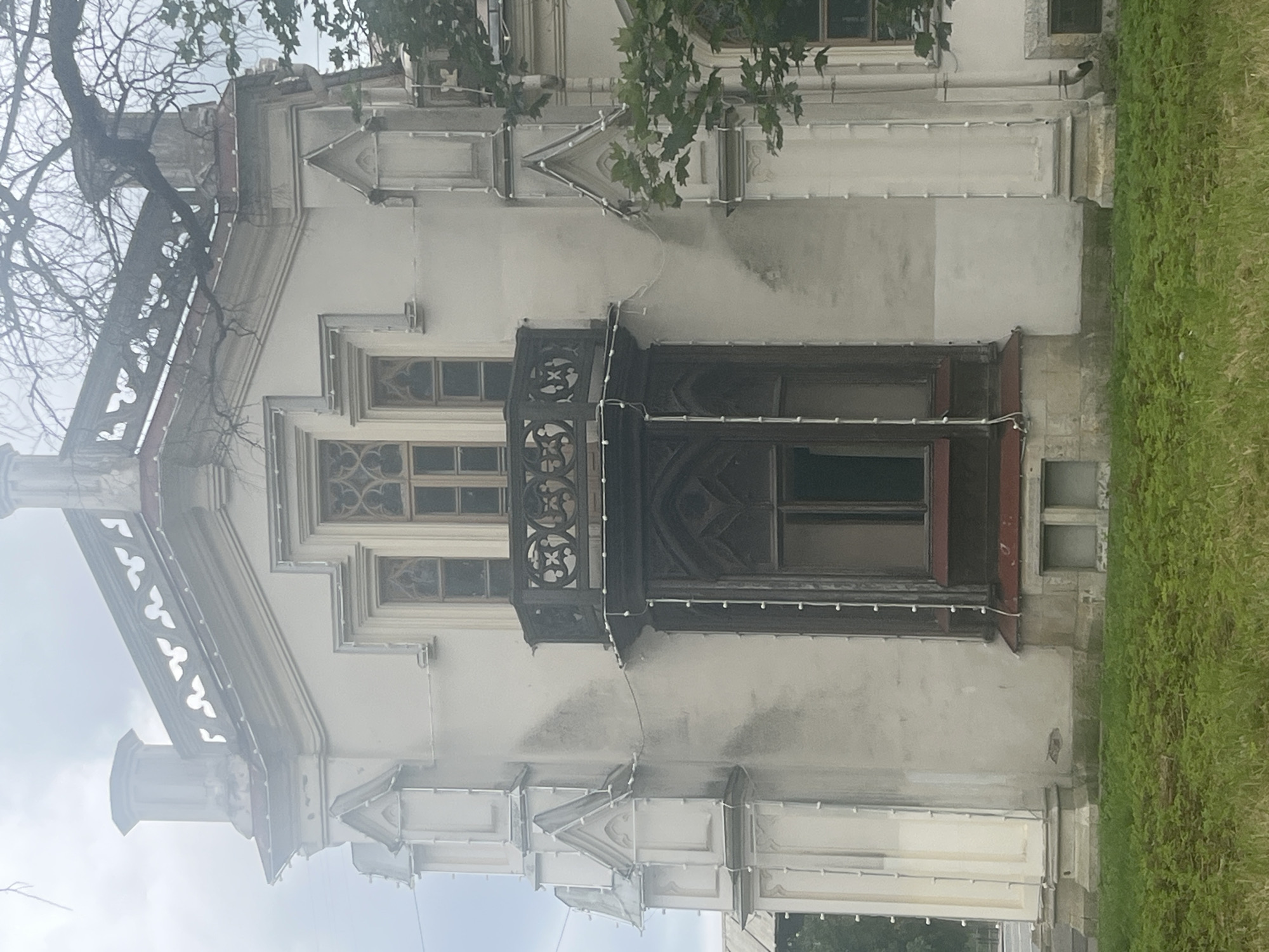 This is a medium shot, eye-level photograph of a stone building with gothic architectural elements, likely a palace or a mansion, located in Peterhof, Russia. The building is predominantly cream-colored with dark wooden window frames and ornate decorative stonework. The facade features a prominent balcony with intricate railings and a bay window beneath it. The roofline is decorated with crenellations. Tall, narrow windows with arched tops are visible on the upper levels. White string lights are draped along the exterior of the balcony and window frames, suggesting recent decoration or illumination. The building is surrounded by a well-maintained green lawn. In the upper right corner, a bare tree branch extends over the building. The sky is overcast and hazy, contributing to a soft, diffused light. There are no people or animals visible in the image.