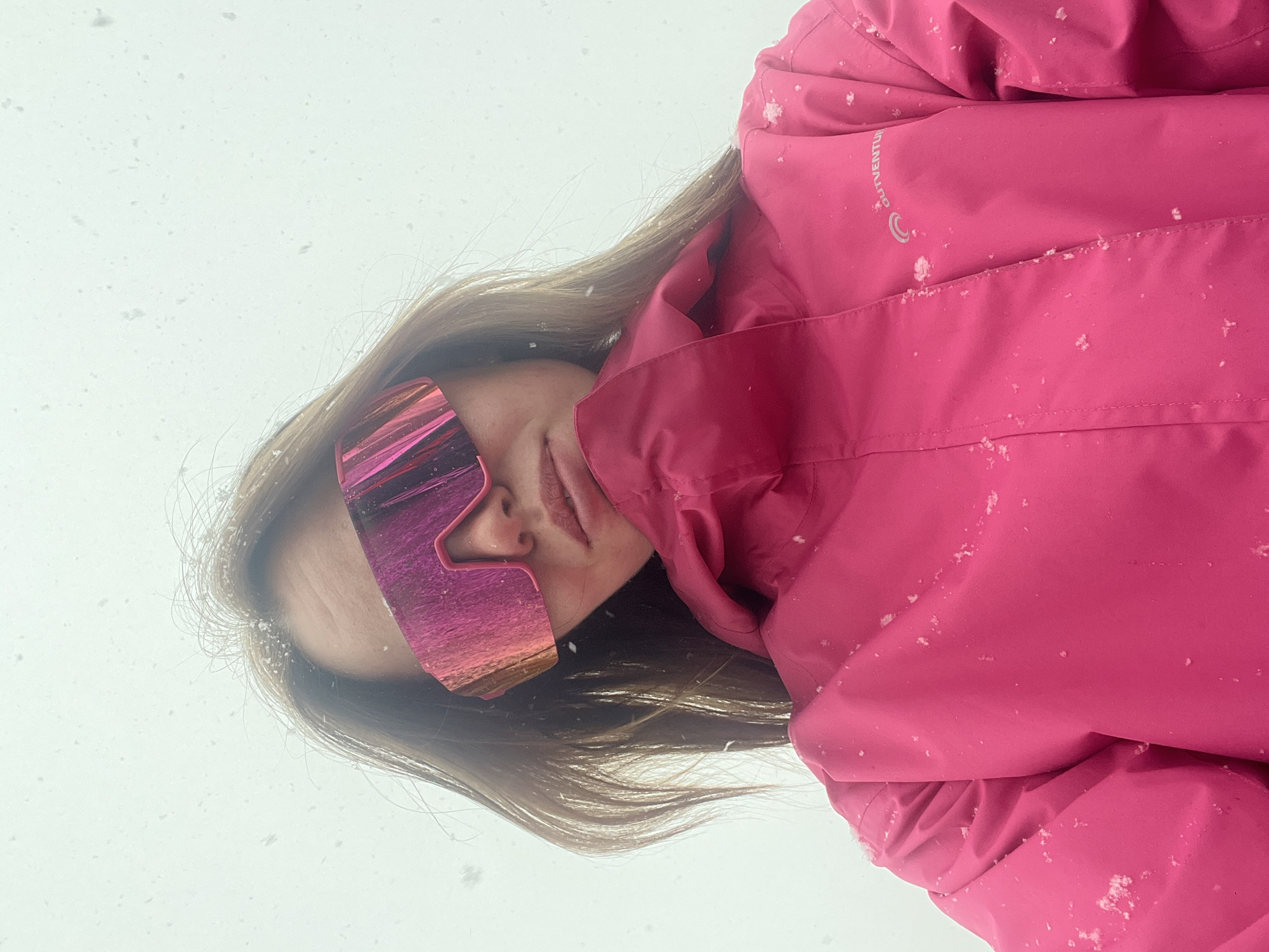 A person wearing pink ski goggles and a pink jacket is shown from a low angle, against a white snowy backdrop. Snowflakes are visible on the jacket and in the air. The location is Kliuchi, Russia, as snow is present in the background.