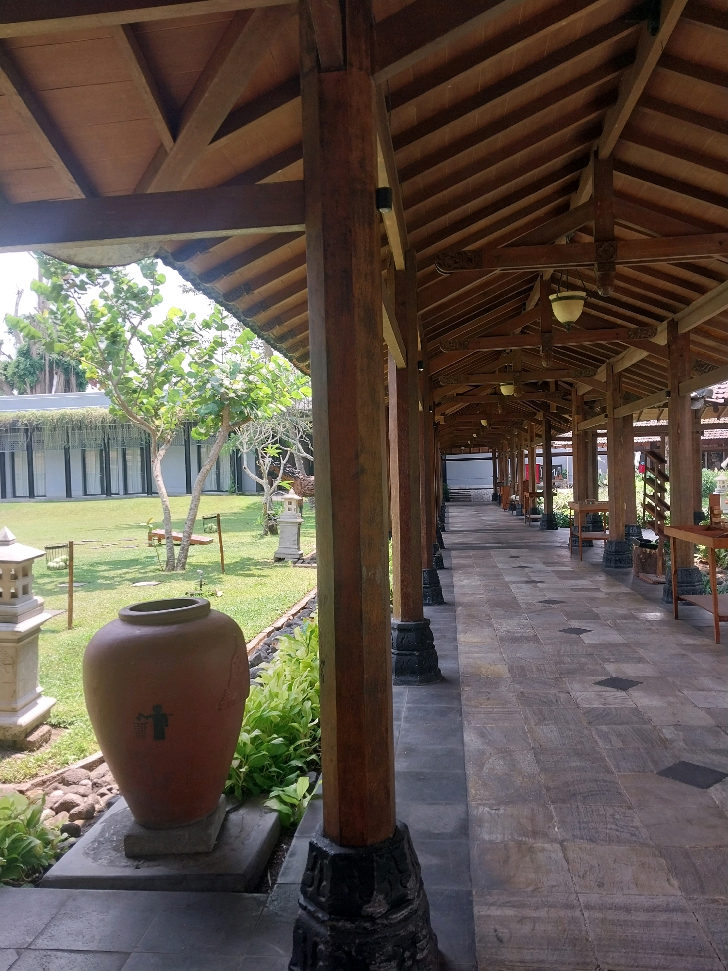 A covered walkway with a wooden roof structure supported by thick wooden pillars leads to a garden area in Ngaglik, Indonesia. The walkway is paved with stone tiles and runs alongside a neatly mowed lawn with several trees and some decorative stone lanterns. In the foreground, a large, reddish-brown ceramic jar, possibly a trash receptacle, sits on a stone base. The walkway extends into the distance, with more pillars and a continuation of the tiled floor. The overall impression is one of a tranquil, outdoor setting with traditional architecture.