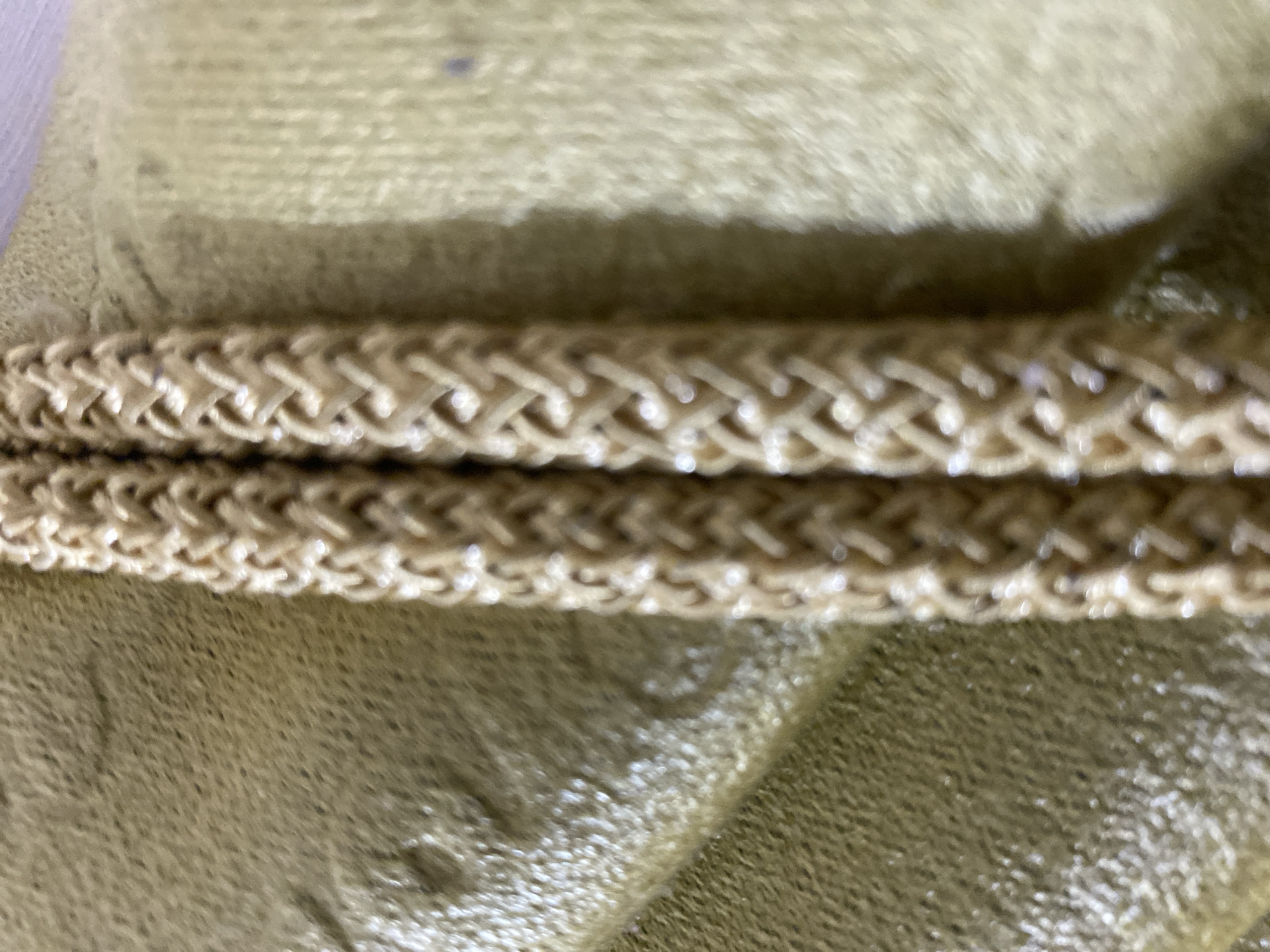 The image shows a close-up, macro shot of two woven, light brown cords lying on a textured, olive-green surface. The cords are parallel to each other and appear to be made of a fibrous material, possibly cotton or a synthetic blend, with a braided pattern. The olive-green surface has a slightly wrinkled or creased appearance and a matte finish, with subtle variations in tone and texture. The lighting is soft, creating gentle highlights on the woven cords and a slight sheen on the green surface. There are no people or discernible signs of the city of Lambaro Kueh, Indonesia, in this image. The focus is entirely on the texture and pattern of the materials.