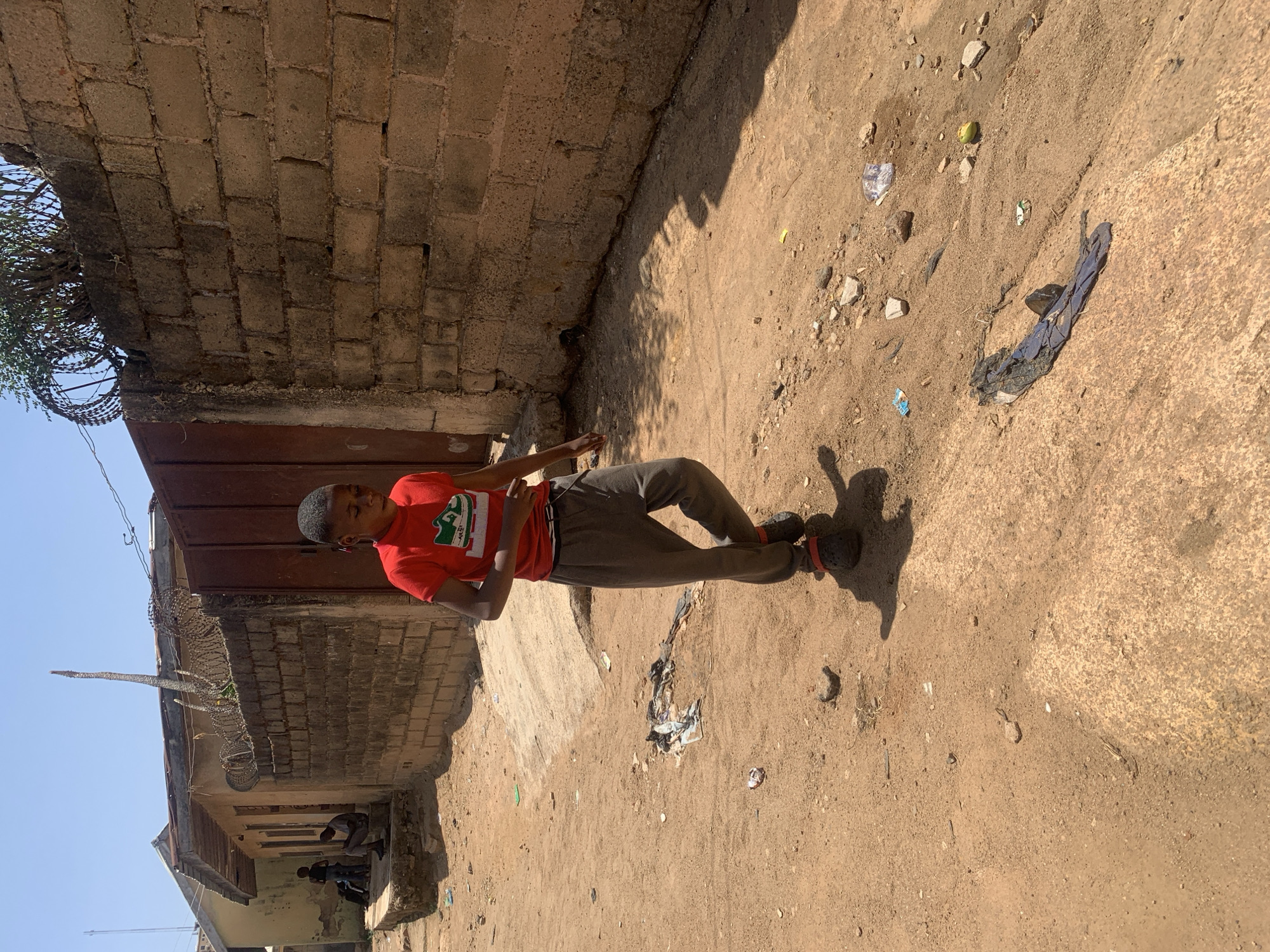 A young person in a red t-shirt and gray sweatpants stands in a sunlit alleyway in what is described as the city of Jos, Nigeria. The alley is narrow, bordered by a brick wall on the left, topped with barbed wire, and a weathered concrete surface littered with trash on the right. The ground of the alley is dirt and sand, also littered with debris. Other people are visible in the background underneath an overhang.
