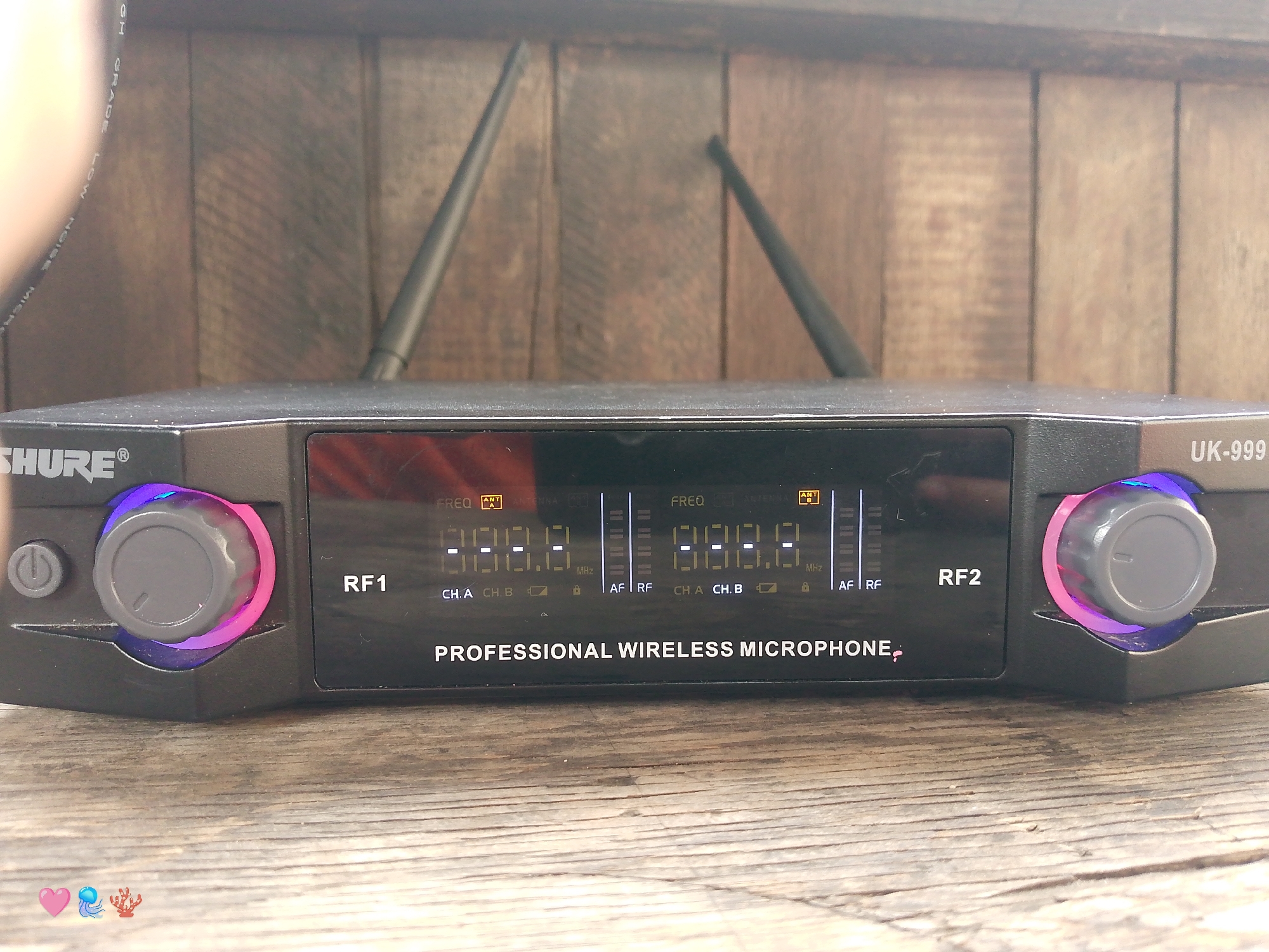 A black Shure UK-999 professional wireless microphone receiver unit is centered in a close-up, eye-level indoor photograph. The device features a central rectangular black display panel showing digital frequency readings (displaying "888.8 MHz" for both RF1 and RF2 channels), "CH.A CH.B" indicators, "ANT A" and "ANT B" antenna status, and vertical signal strength bars for "AF" (Audio Frequency) and "RF" (Radio Frequency). To the left of the display, a power button and a grey rotary knob with a purple and pink illuminated base are visible. A matching grey rotary knob with a similar illuminated base is on the right side of the display. Two black antennae extend diagonally upwards from the rear of the unit. The receiver rests on a light brown wooden surface with horizontal planks. The background consists of a darker brown wooden wall or paneling with vertical planks. A partial, out-of-focus light-colored object, possibly a finger or hand, is visible in the upper left, partially obscuring a black cable with faint white text including "GRADE LOW NOISE MICRO". Three digital emojis (pink heart, blue jellyfish, orange coral) are superimposed in the bottom left corner of the image. The image does not contain any visual information to determine its location in the city of Pedro González, Venezuela.