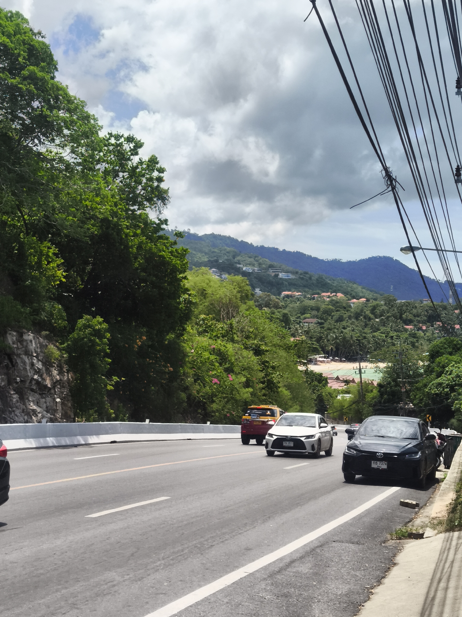 The image depicts a scenic view of a road in Ko Samui District, Thailand, likely a coastal area given the distant beach and turquoise water. The road is paved and appears to be a two-lane highway, with white lines marking the lanes and a yellow line dividing the directions of traffic. Several vehicles are visible on the road: a black sedan in the foreground, a white sedan and a yellow taxi further ahead, and the rear of a red vehicle just beyond. The road is bordered on the left by a steep, rocky incline covered in lush green vegetation. On the right, a concrete sidewalk runs alongside the road, with a curb and a patch of grass and dirt. Overhead, a tangle of electrical wires crisscrosses the sky, supported by poles. In the background, rolling hills covered in dense tropical foliage are dotted with buildings, suggesting a residential or resort area. A glimpse of a beach with umbrellas and buildings can be seen between the trees on the far side of the bay. The sky is overcast with dramatic clouds, but patches of blue are visible, indicating diffused sunlight. The overall impression is a tranquil tropical landscape with a functional roadway.