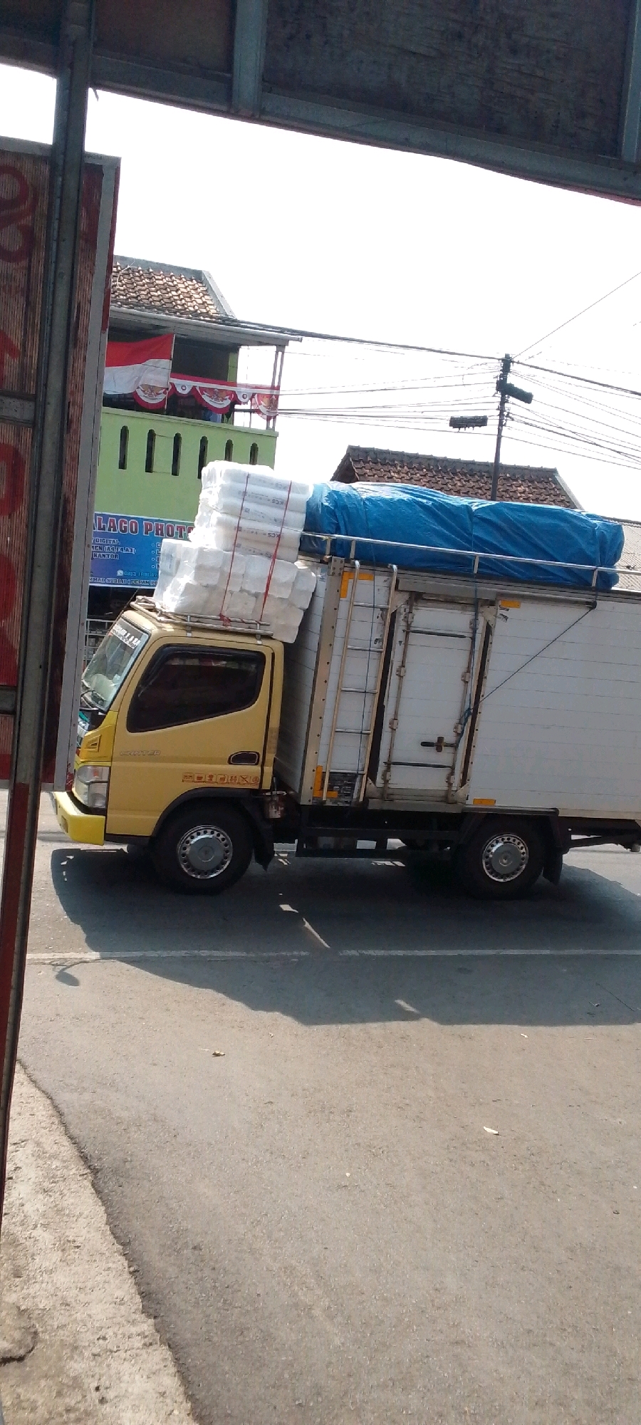 A yellow truck is parked on the side of a street in Cilaku, Indonesia. The truck is loaded with white rectangular packages stacked high on the roof and covered with a blue tarp. The truck has a white cargo box with a ladder attached to the side. In the background, there is a building with a tiled roof decorated with Indonesian flags. The street is paved and there are shadows cast on the ground. The overall scene suggests a commercial or logistical operation taking place in an urban environment.