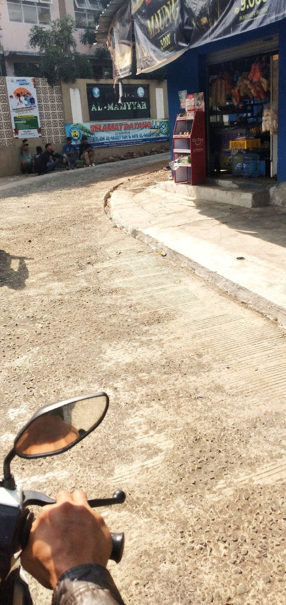 A first-person perspective shot shows a hand on a motorcycle handlebar, with the vehicle's side mirror reflecting an orange-toned view of the rider's forearm. The scene is an outdoor environment with a paved road made of rough gravel and concrete. In the background, a group of five men are seated on the ground next to a low wall. Behind them, a building with a sign that reads "AL-MAMY'YAH" is visible. A red display stand with various items is placed in front of a blue building which appears to be a shop, with goods hanging inside. The image captures a street in Cilaku, Indonesia.