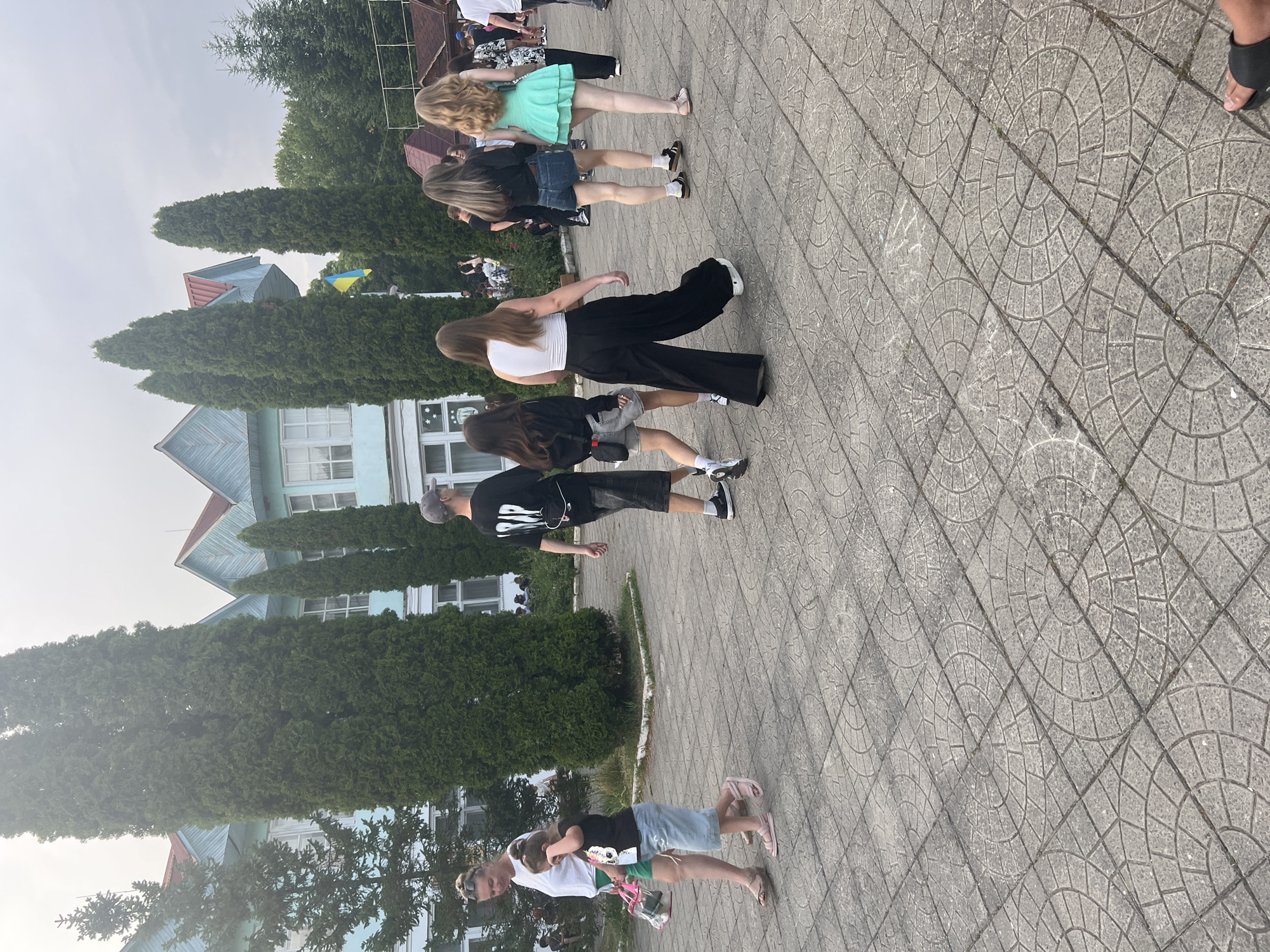 A group of people are walking on a paved area in front of a light blue building with a red roof. The building has several windows and is surrounded by tall green trees. A Ukrainian flag is visible on a flagpole to the right of the building. The ground is paved with gray tiles that have a circular pattern. A woman in a white shirt and green shorts is walking with a young girl in a black t-shirt and denim shorts. A man in a black t-shirt and shorts is walking behind them. Several other people are walking in the background.