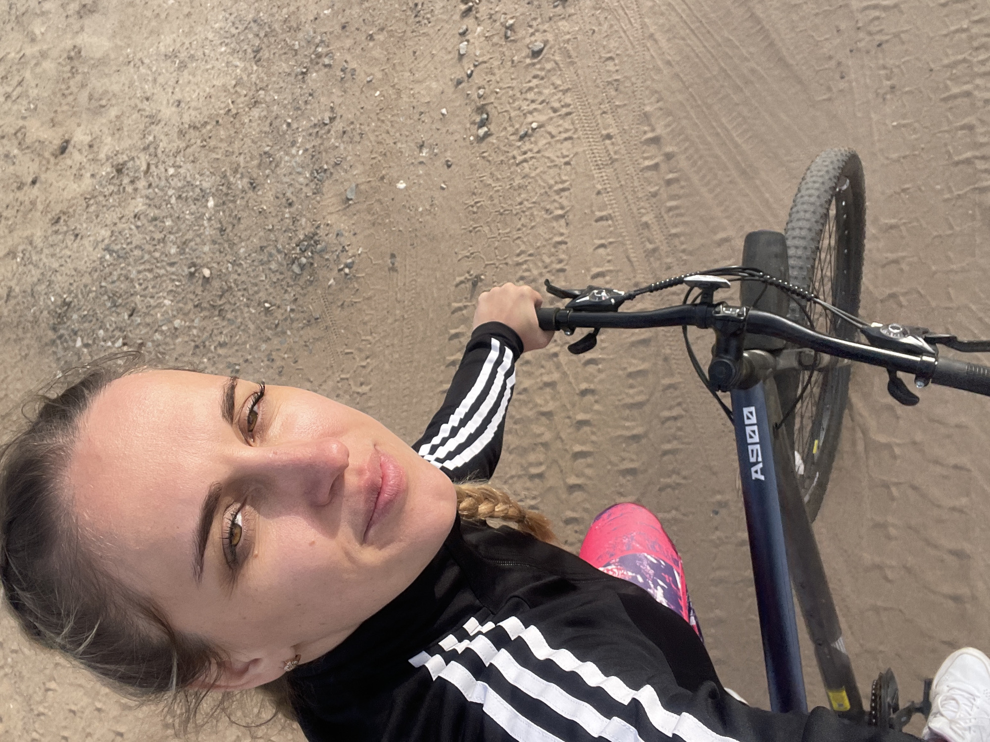 A woman is taking a selfie while riding a bicycle on a sandy path. She is wearing a black Adidas tracksuit with white stripes and colorful leggings. The bicycle is a dark color with the model number "A900" visible. The ground is sandy with tire tracks. The location is Chaikovsky, Russia.