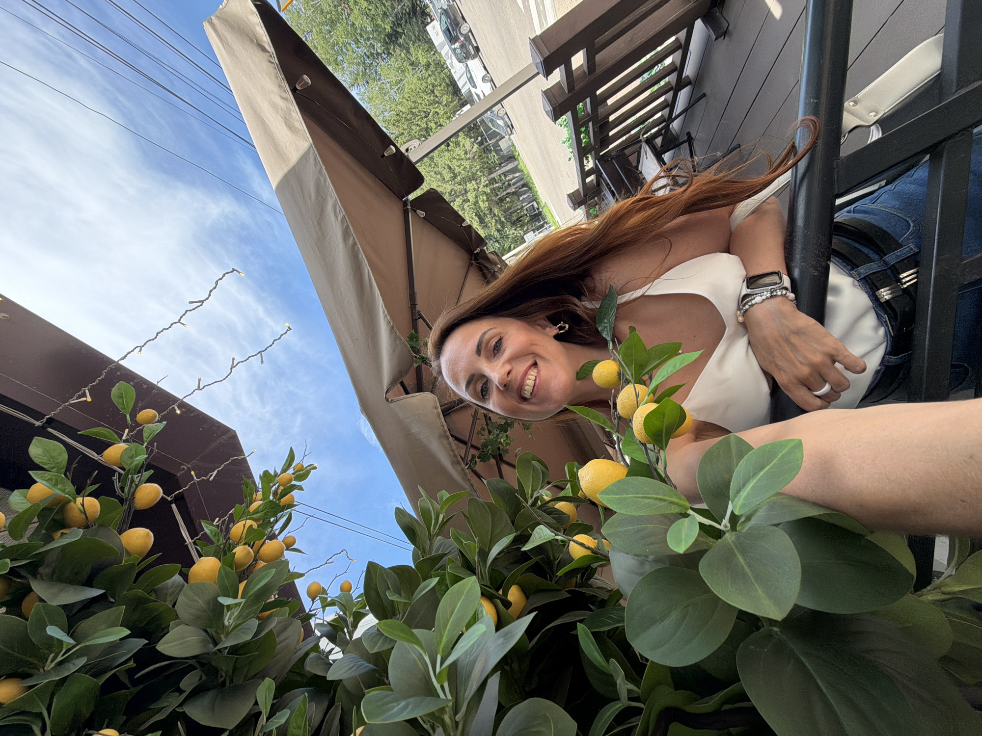 A woman with long, auburn hair is posing for a selfie outdoors, with a backdrop of artificial lemon branches, a patio umbrella, and a bright blue sky with some clouds. She is wearing a white tank top, blue jeans, and a black belt. She has a bracelet on her left wrist and a watch on her right wrist. In the background, there are trees, a parking lot with cars, and a wooden railing. The scene appears to be at a cafe or restaurant with outdoor seating. The woman is smiling and looking directly at the camera. The location is specified as Birobidzhan, Russia.