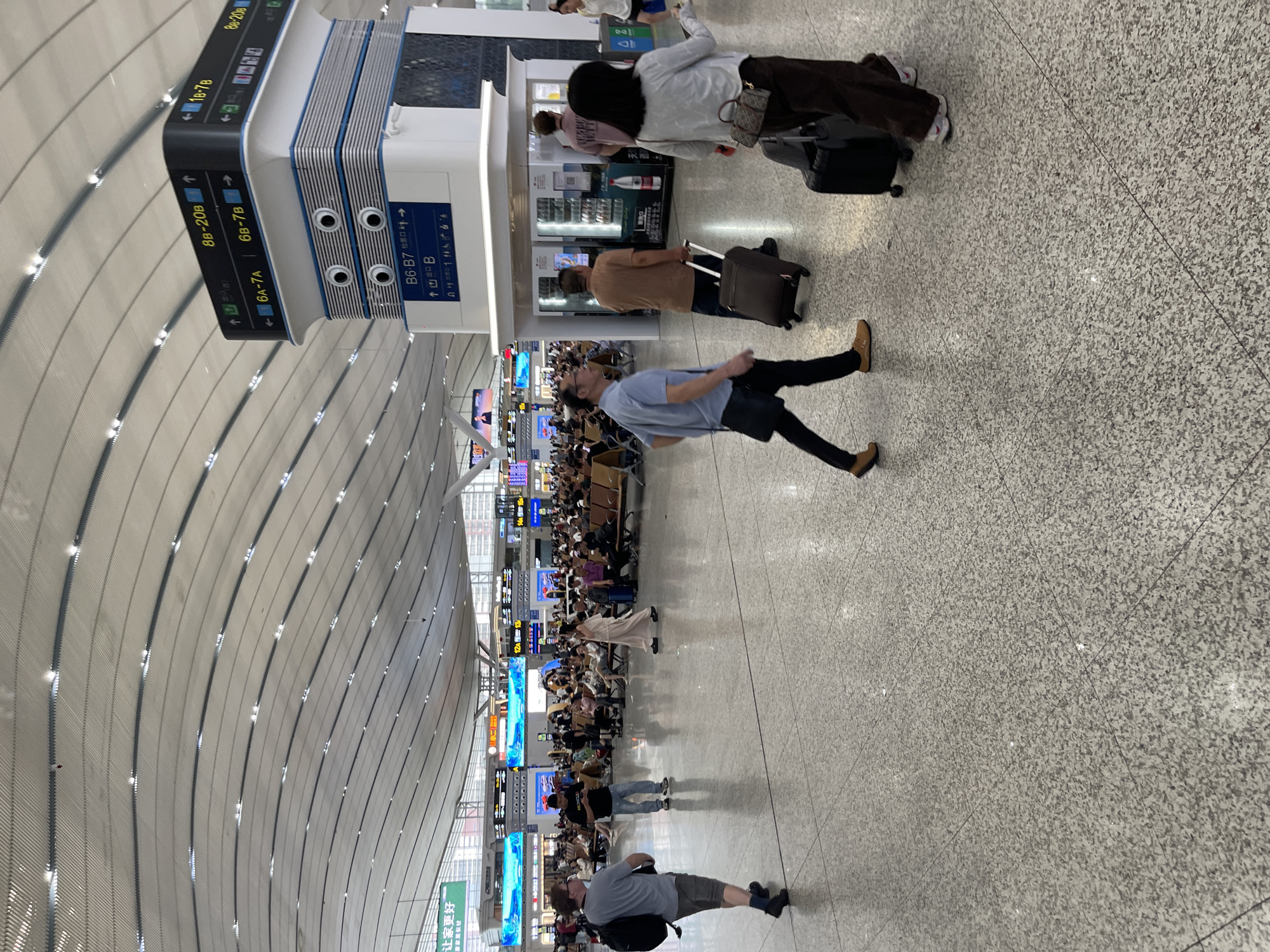 The image depicts the interior of a modern transportation hub, likely an airport or a high-speed train station, in Longhua District, China. The architecture features a sweeping, curved ceiling with integrated lighting, creating a spacious and illuminated environment. In the foreground, several individuals are visible walking across a polished, speckled floor, some with luggage. To the right, a tall, multi-sided signage tower displays directional information in both Chinese characters and Arabic numerals, indicating gates and platforms such as "8B-20B", "6A-7A", and "B6-B7". In the middle ground, a large waiting area is populated with numerous people seated on rows of benches, facing digital display boards that likely show schedules or travel information. Further in the background, more seating areas and information displays are discernible, along with glimpses of the exterior through large windows. The overall scene conveys a sense of organized transit activity.