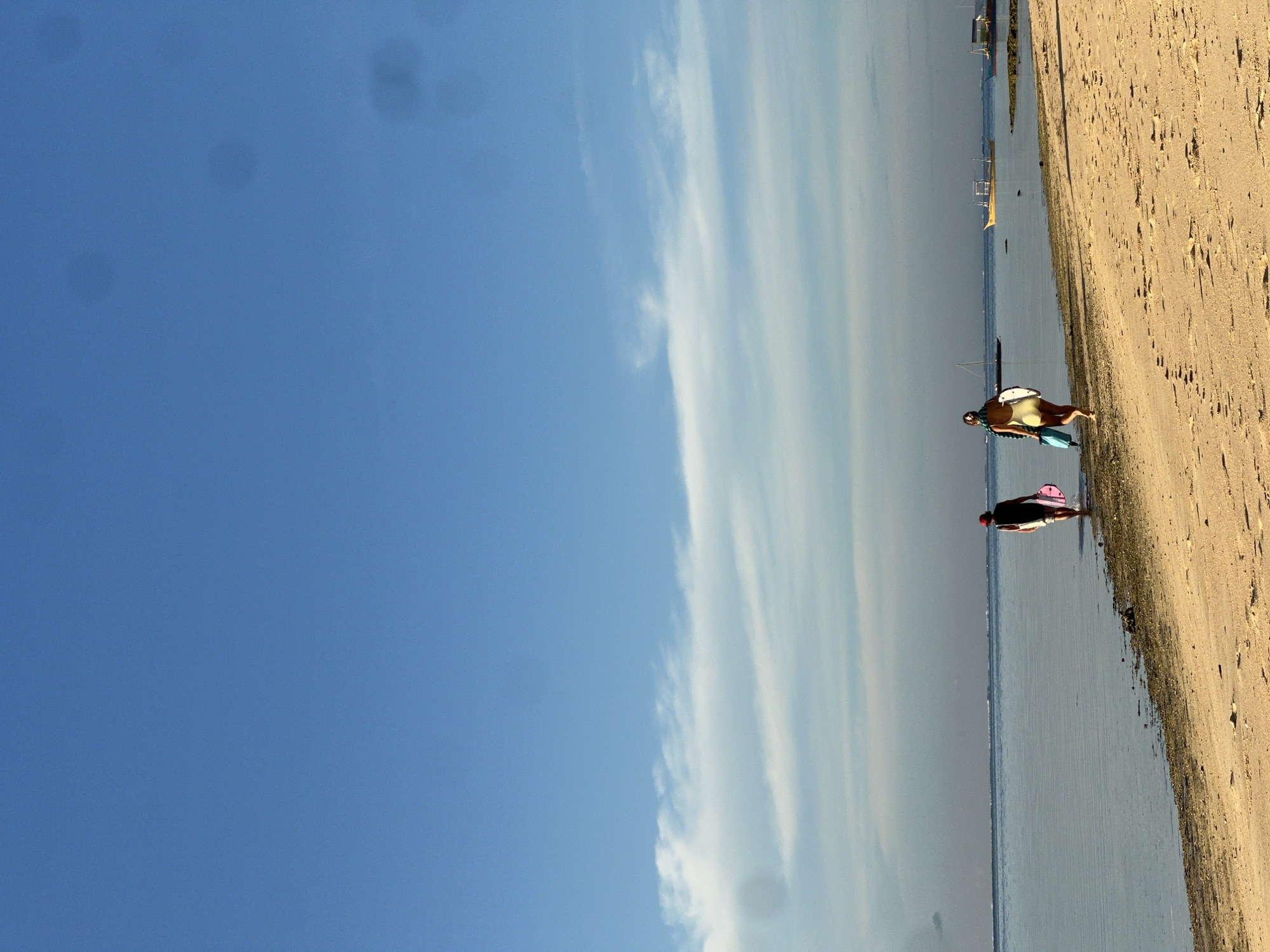 Two individuals are walking on a sandy beach adjacent to a calm body of water. The person on the left is wearing a dark top and white shorts, carrying a pink surfboard. The person on the right is wearing a light-colored swimsuit and carrying a white surfboard and a blue bag. The sun is low in the sky, casting a warm glow on the beach and illuminating thin, wispy clouds. Several small boats are visible in the distance on the water. The scene appears to be a tranquil morning or late afternoon on a tropical beach, likely in the vicinity of General Luna, Siargao, Philippines, known for its surfing spots.