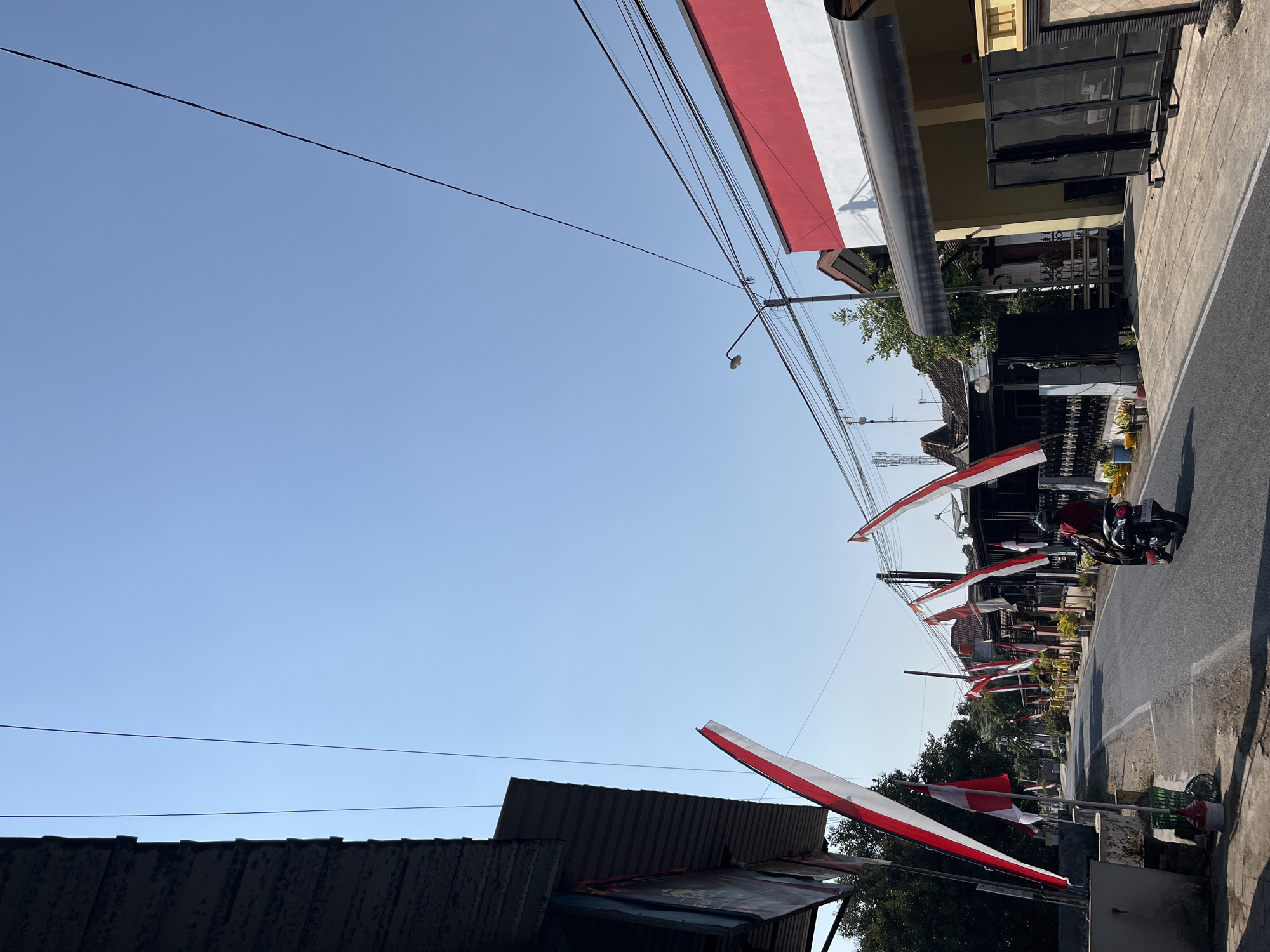 The image depicts a street scene in Margorejo, Indonesia, on a clear, sunny day. The road is lined with houses adorned with numerous Indonesian flags, predominantly red and white, hung on poles and stretched across the street. These flags suggest a celebration or commemoration. A person is riding a motorcycle away from the camera down the asphalt road. The architecture visible includes houses with tiled roofs and some modern structures with awnings and fences. Utility wires are strung across the sky, and a street lamp is visible. The overall environment appears to be a residential area with a sense of community and national pride.