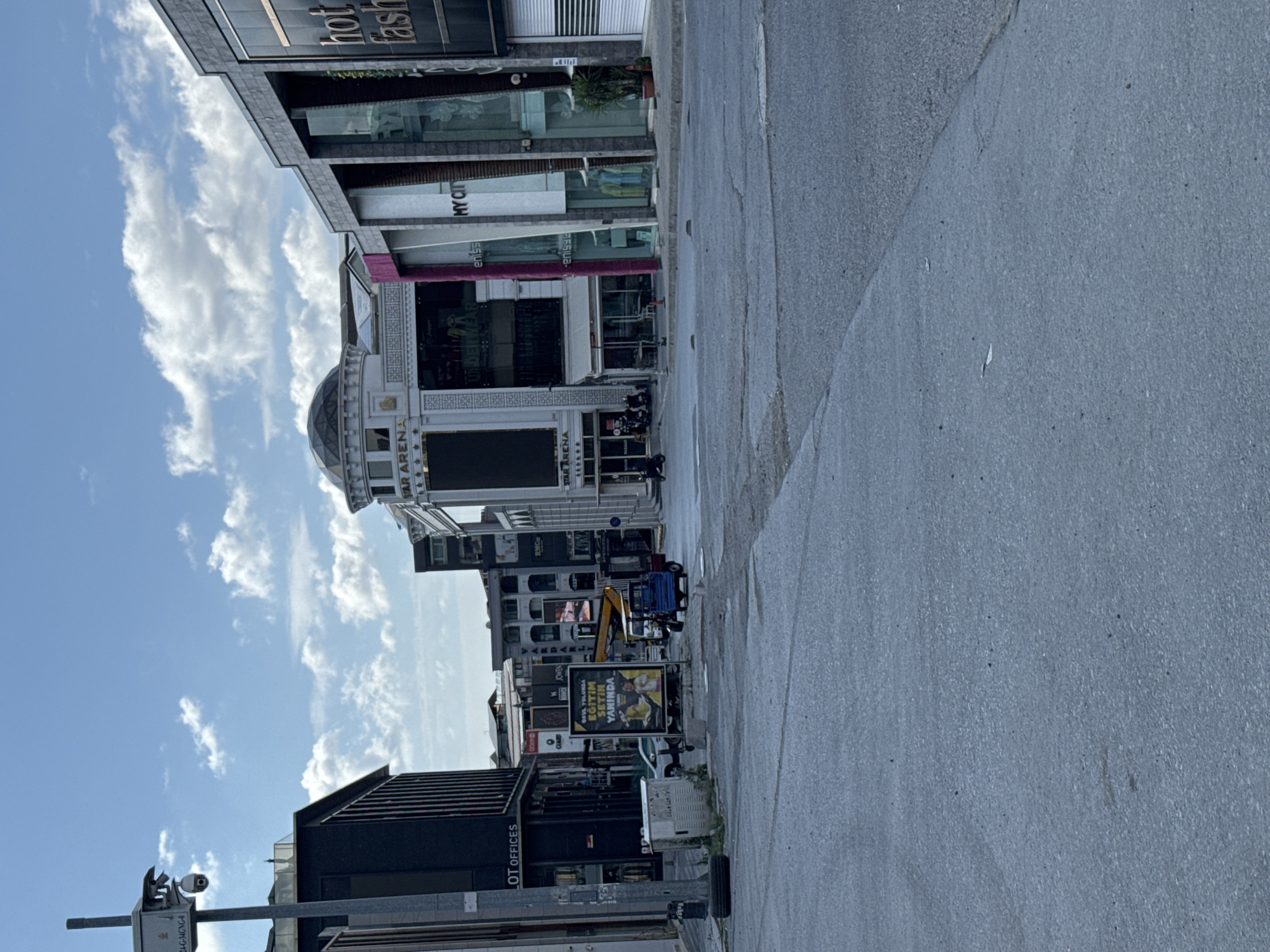 The image depicts an outdoor scene at street level, showcasing a commercial area in Gungoren, Turkey. The perspective is from a road, looking towards a row of buildings with various storefronts. The sky above is partly cloudy, with visible cumulus clouds against a blue backdrop.
In the foreground, the asphalt road is visible, appearing somewhat worn and showing patches of discoloration. The left side of the image features a tall, dark building with the sign "LOT OFFICES" visible. To the right of this building, a streetlamp with a security camera mounted on it stands. Further into the scene, several other buildings line the street, exhibiting different architectural styles and facade designs. One building has a prominent domed roof and a large digital screen displaying text and images, with the words "STAR ARENA" clearly visible. Adjacent to this, other storefronts display signage such as "UNDERWEAR", "eniss", and "MY CITY". The building on the far right has a modern, gray facade with large glass windows and a closed shutter.
Several objects are present on the street. A blue motorized three-wheeled vehicle is parked in the middle ground. A white vehicle is also visible, as is a large yellow construction vehicle partially obscured. A billboard with Turkish text and images is positioned on the left side of the street. There are also a few motorcycles parked near the storefronts. Some planters with greenery are placed outside some of the establishments. A white electrical box is situated on the left side of the road, next to a discarded tire.
The scene appears to be devoid of significant human activity, with only a few individuals visible in the distance, either walking or tending to something on one of the buildings. The overall impression is a quiet urban environment under daylight.