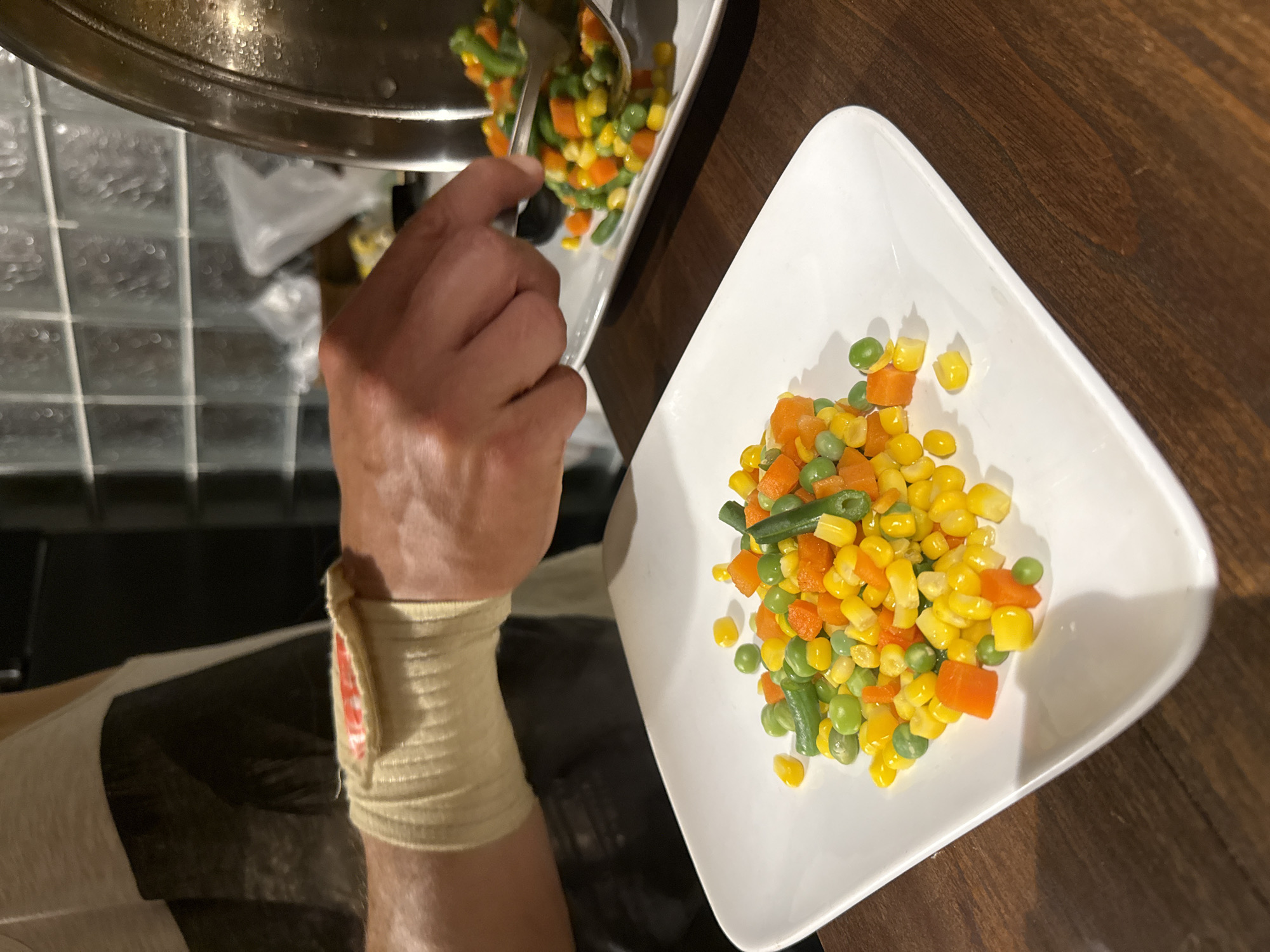 In this close-up, high-angle shot, a person's hand, wrapped in a beige bandage, is seen scooping mixed vegetables from a pot into a white bowl. The vegetables consist of corn kernels, diced carrots, green beans, and peas. The pot is metallic and appears to be steaming, suggesting the vegetables are freshly cooked. The white bowl is irregularly shaped and partially filled with the colorful mixture. The background shows a dark, wooden surface and a tiled area that appears to be a countertop. The lighting is dim, hinting at an indoor setting, possibly during the evening or in a poorly lit kitchen. The focus is on the action of serving the food, with the hand and the vegetables being the primary subjects. No visible text is present in the image.