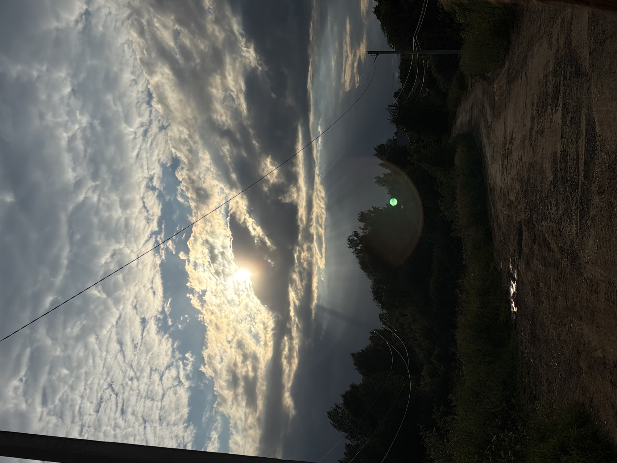 The image depicts a rural scene in the vicinity of Фоки, Russia. The primary subjects are the sky and the surrounding landscape. The sky is filled with a dramatic display of clouds, with the sun breaking through in the center, casting rays of light. The clouds appear textured and varied, ranging from bright white to dark grey, suggesting dynamic weather conditions.
The setting includes a dirt road or path on the right side of the frame, lined by dark green trees and vegetation. Power lines are visible, stretching across the sky, indicating the presence of infrastructure in this rural area. A noticeable green lens flare is present in the upper right quadrant of the sky, caused by the sun's light. The overall lighting suggests it is either sunrise or sunset, with the low angle of the sun creating long shadows and highlighting the cloud formations.
No people are visible in the image. The scene conveys a sense of natural beauty and tranquility, with the dramatic sky as the focal point. The details of the dirt road and the lush greenery suggest a natural, undeveloped environment. The presence of power lines provides a subtle hint of human habitation. The time of day is likely late afternoon or early evening, given the quality of light and the direction of the sun. The weather appears to be partly cloudy.