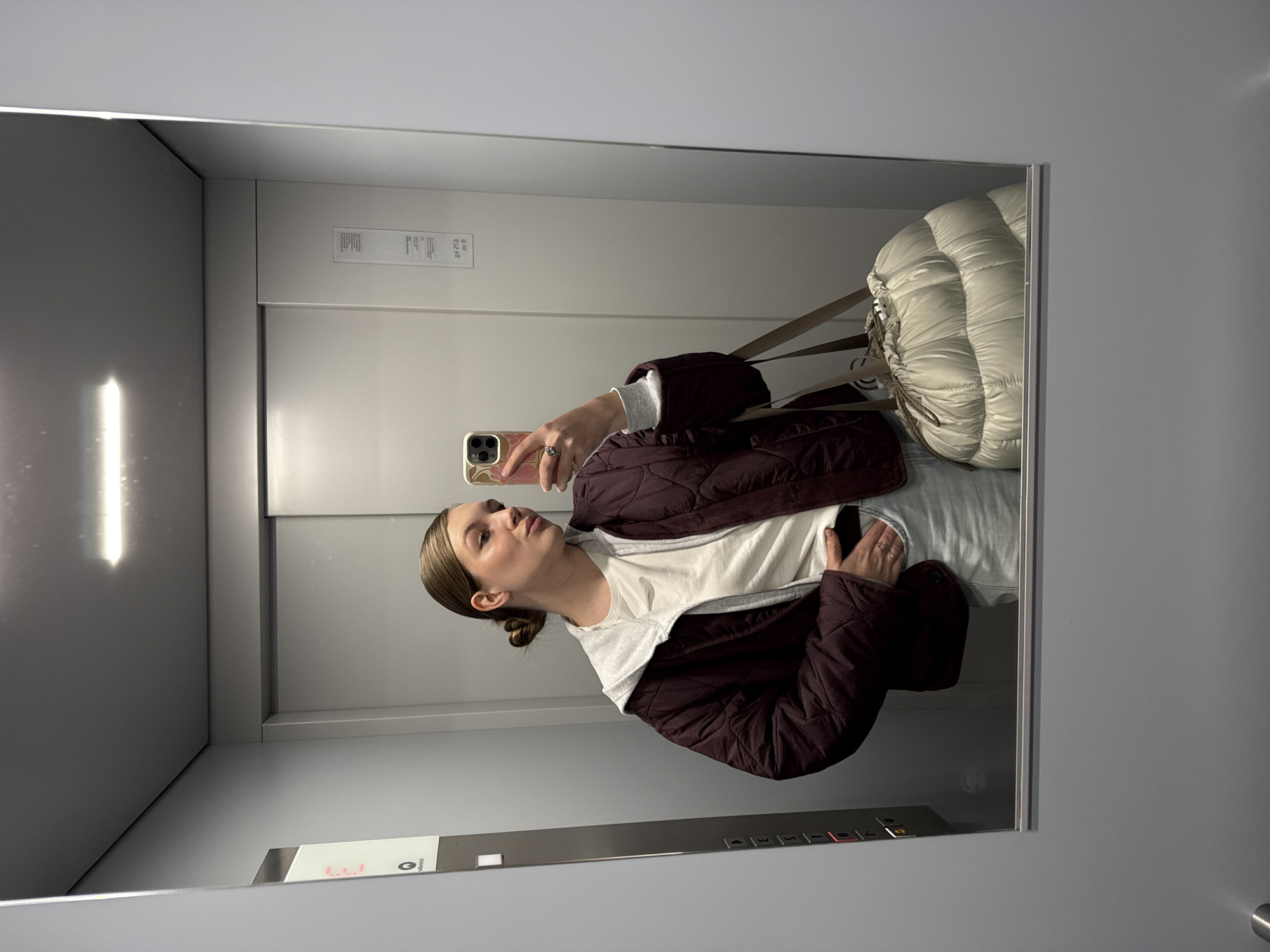 A woman is taking a selfie in a mirror located inside an elevator. She is wearing a burgundy quilted jacket over a white hooded sweatshirt and gray pants. A large, light-colored bag with straps is also visible in the mirror, positioned behind her. The elevator interior is predominantly gray with a control panel showing floor numbers. The location cannot be determined.