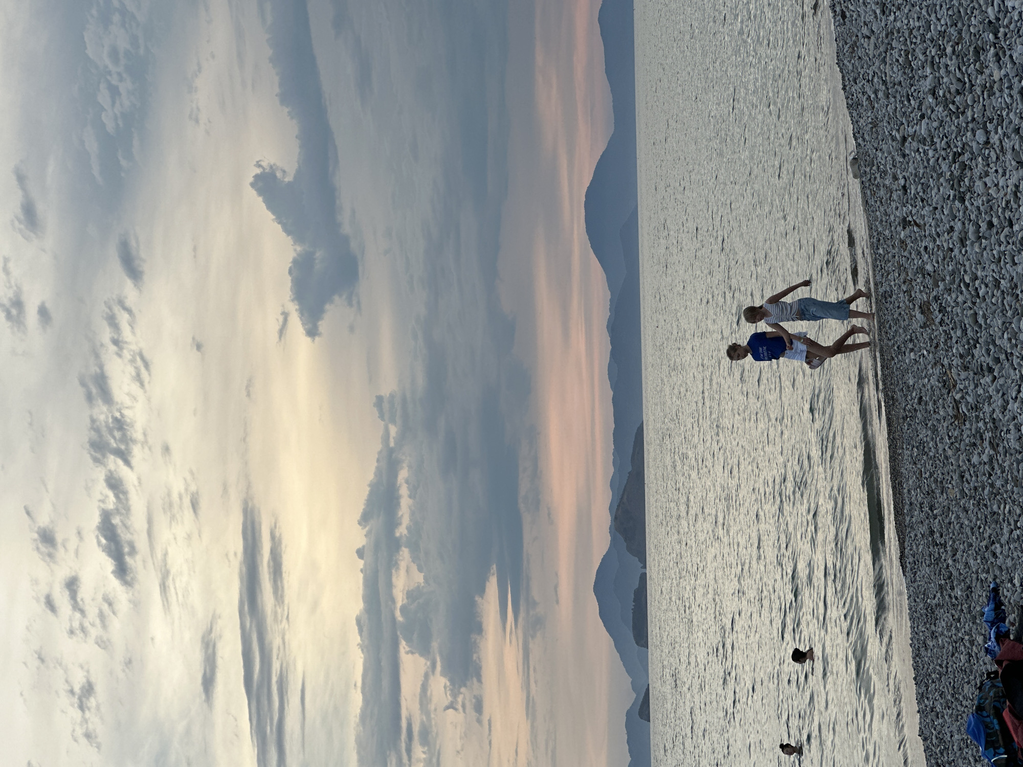 The photograph captures a coastal scene in Fethiye, Turkey, at what appears to be sunset or dusk. The horizon line is demarcated by layered mountains, which fade into the distance under a sky filled with wispy and striated clouds. The light is soft, with hints of pink and gold peeking through the cloud cover.
The right side of the image is dominated by a beach made of pebbles or small rocks. Two people, likely children, are walking along the water's edge. One is wearing a blue shirt, and the other appears to have on a striped top.
A few dark objects are visible in the water, potentially birds or other items floating on the surface. In the lower right corner of the frame, there are a few piles of what look like clothing or bags, indicating that the beach is being used by people.