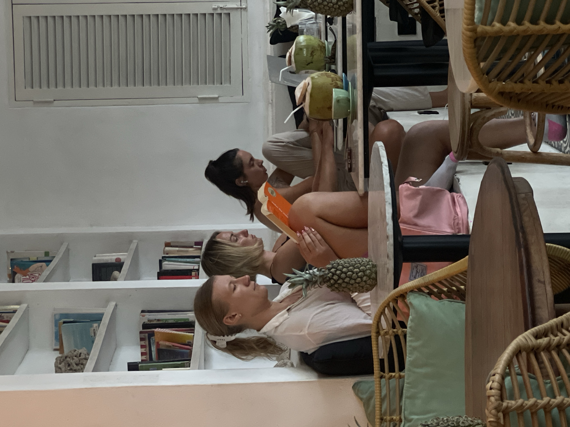 The image depicts the interior of a building, possibly a café or a shared workspace, in North Kuta, Indonesia. The foreground shows a seated woman with light hair in a white shirt and black cushions. She is holding a pineapple and has a bookshelf behind her. Further back, another woman reads a book. Near her is a person wearing a light-colored garment with pink socks. In front of them are coconut drinks placed on a table. The seating consists of rattan chairs with pale green cushions.